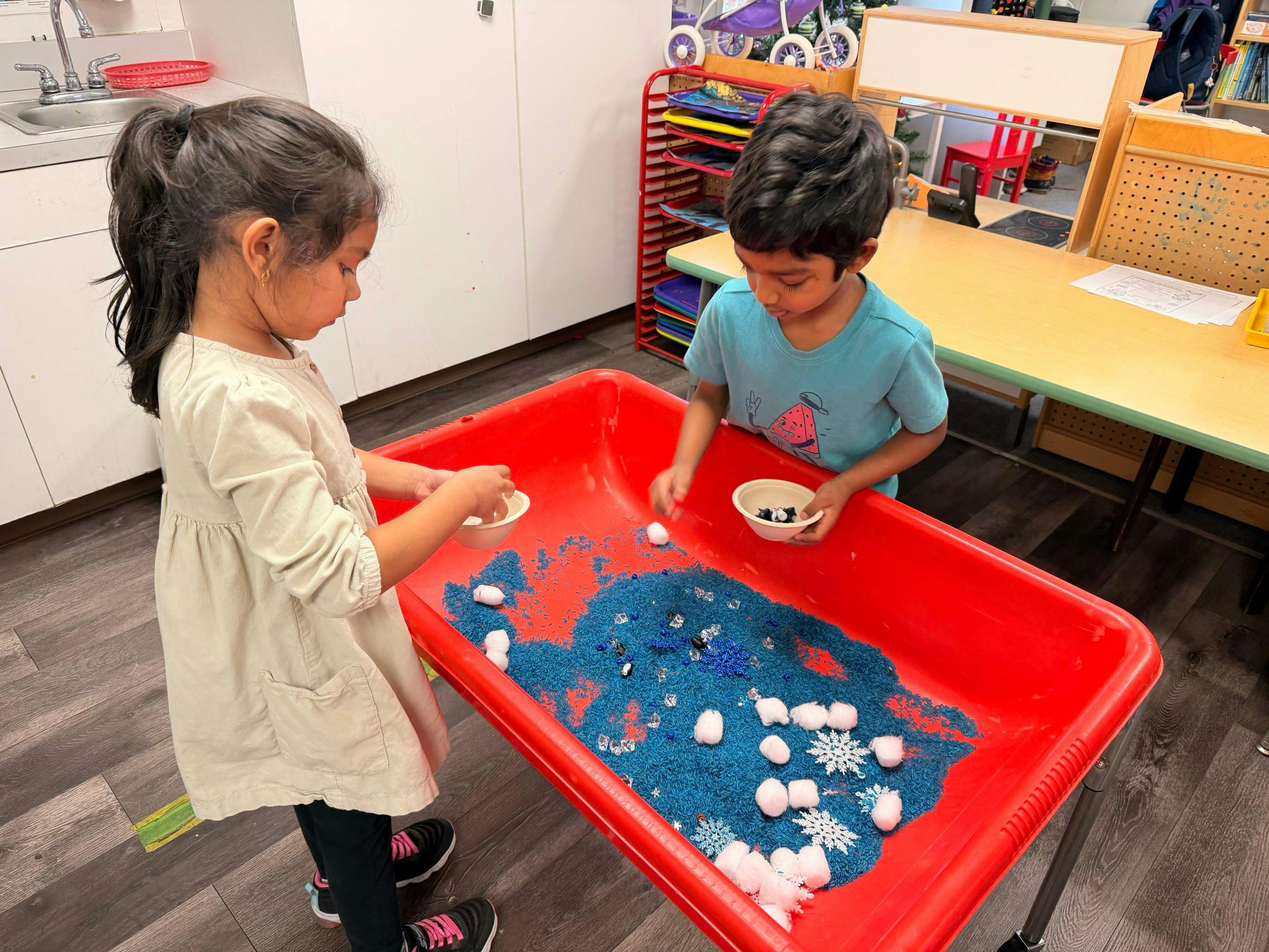 Two children playing with blue rice, cotton balls, and snowflake decorations in a red bin