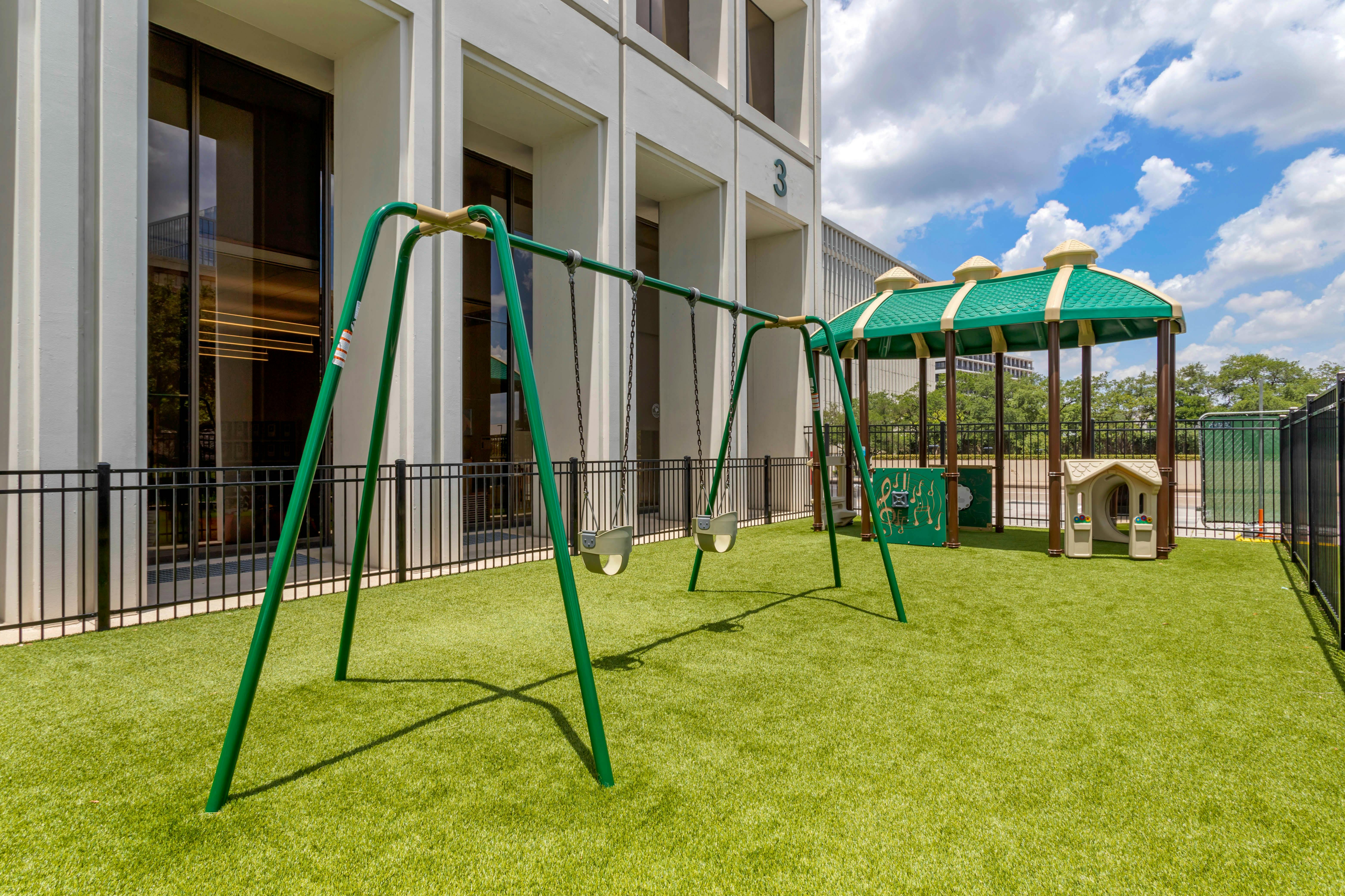 A swing set and small covered play structure on artificial grass in front of a building