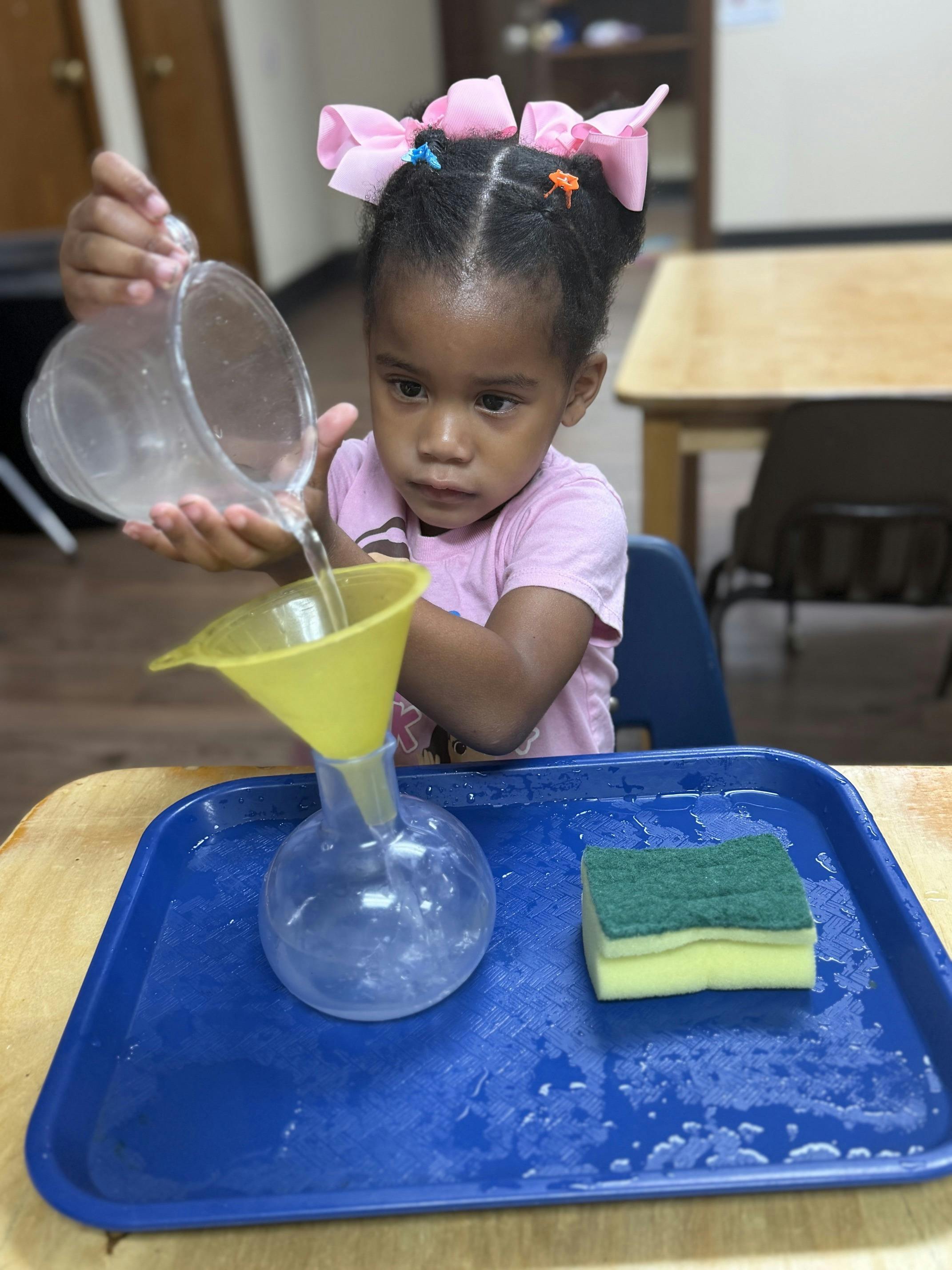 Child pouring water into a flask using a funnel on a tray