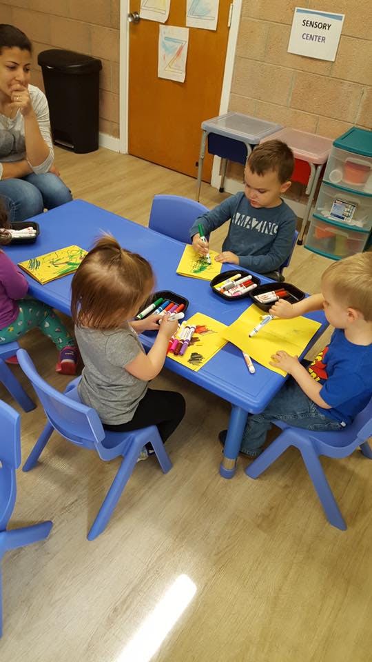 Children drawing at a blue table in a classroom with an adult nearby