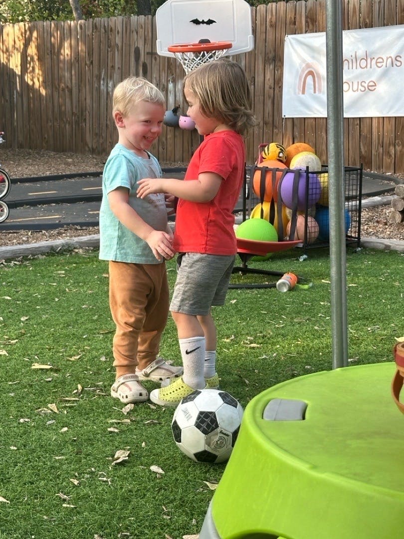Two children standing and facing each other on artificial grass near a soccer ball