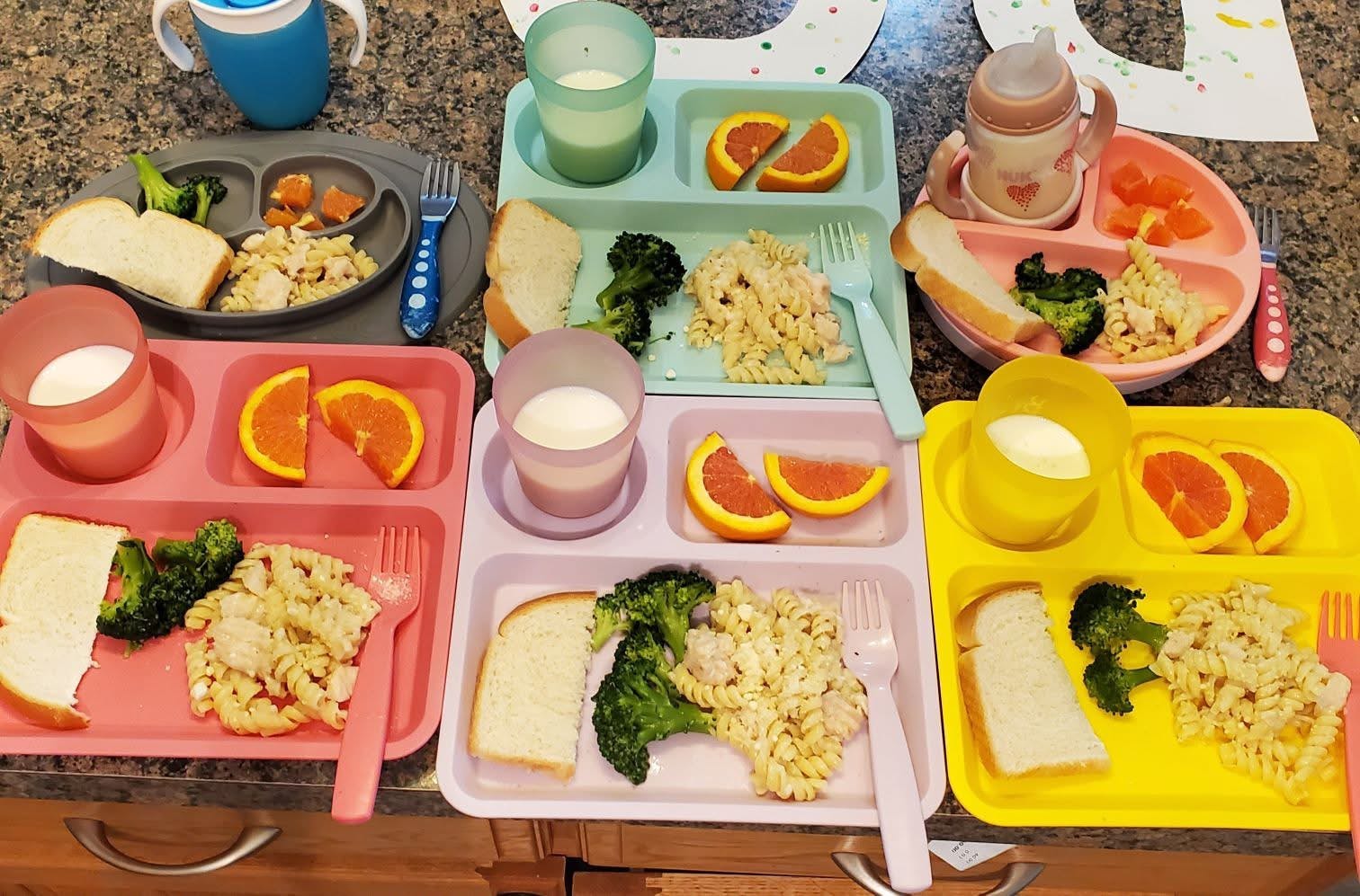 Several divided plates containing pasta, broccoli, bread, and orange slices