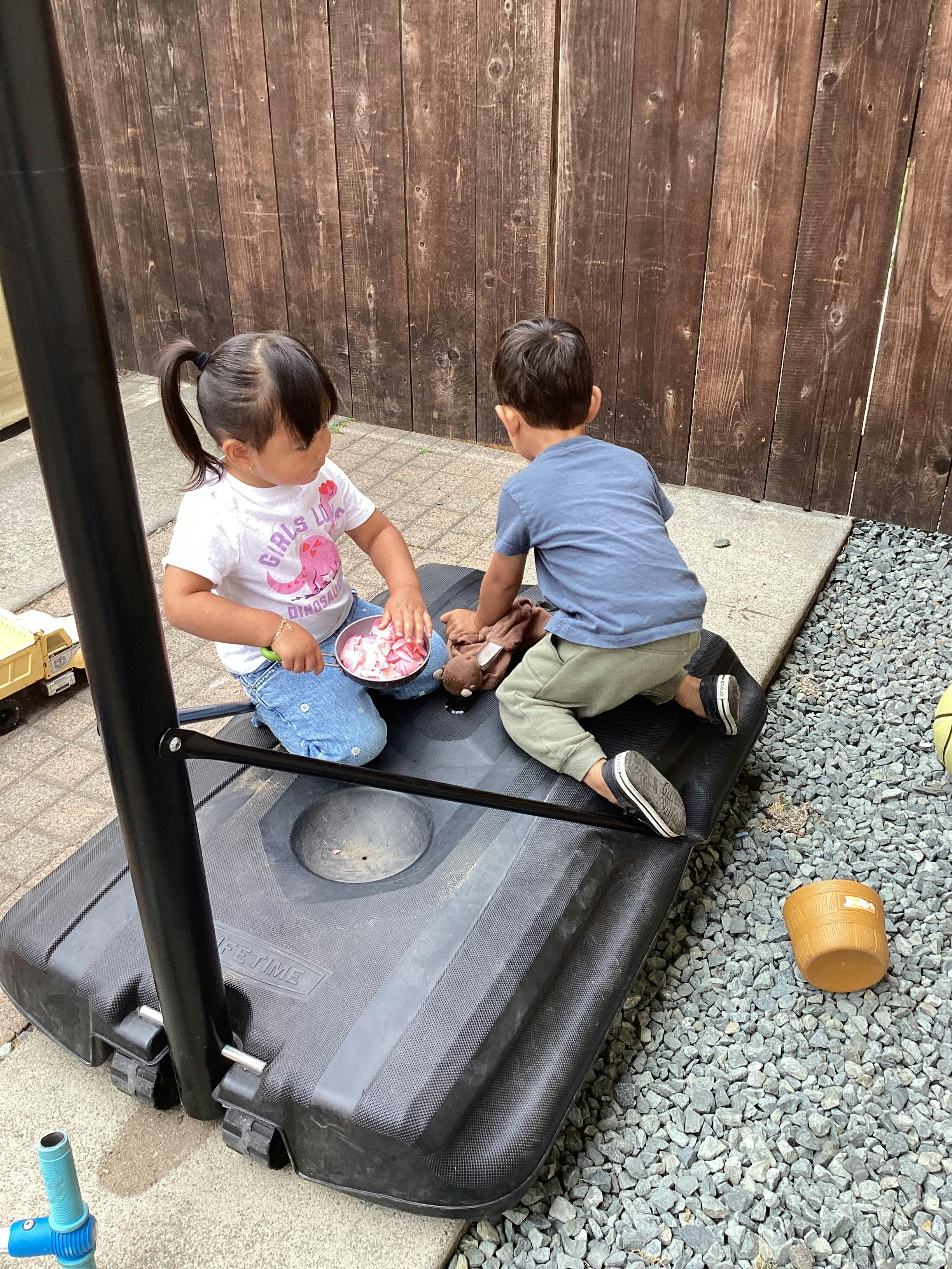 Two children playing with a small pan on a basketball hoop base outdoors