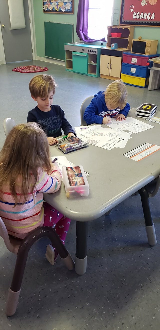 Three children sitting at a table, two writing and one looking downcast
