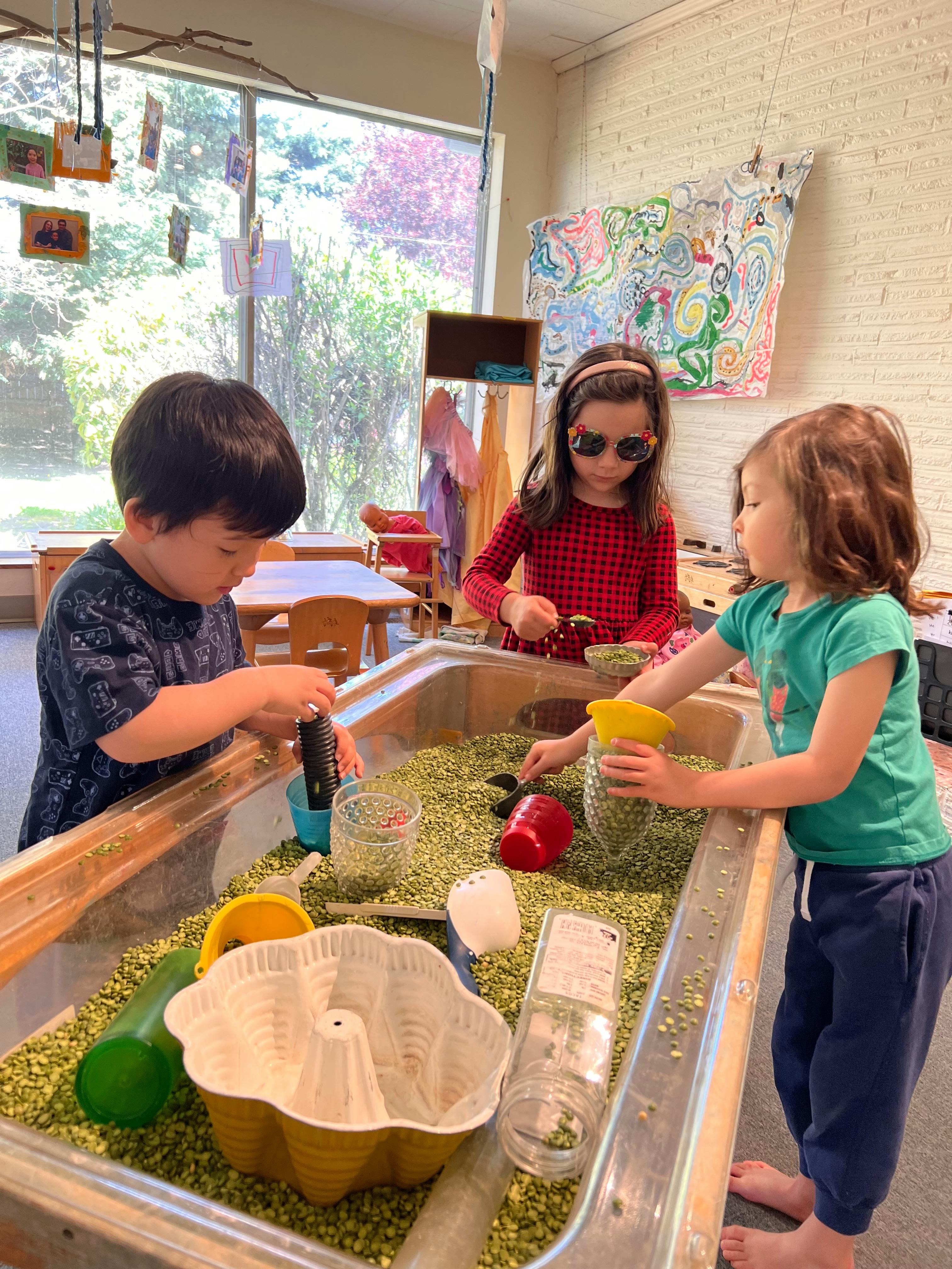 Three children playing at a sensory table filled with green peas