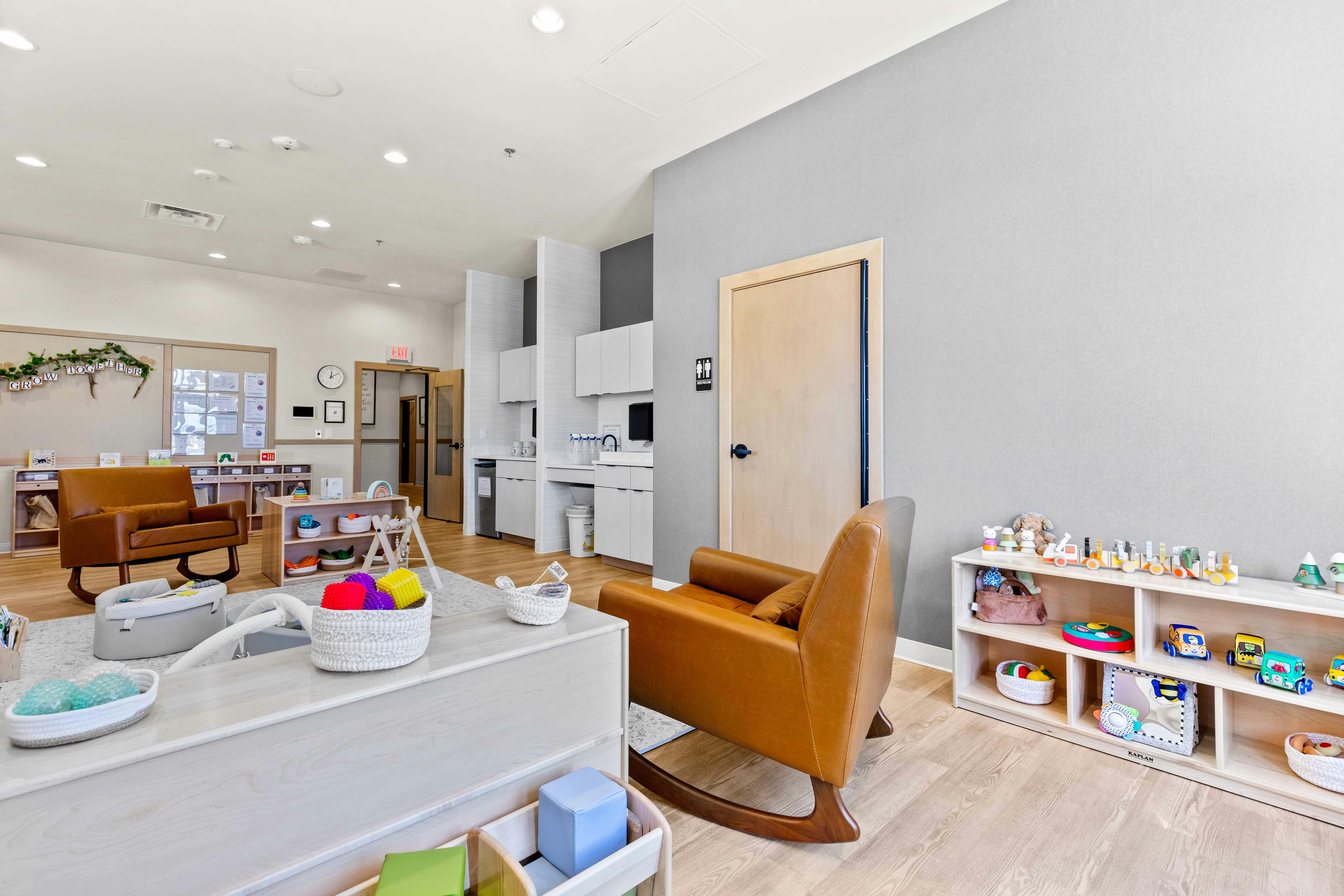 A room with a brown rocking chair, a white shelf with toys, and a small kitchen area