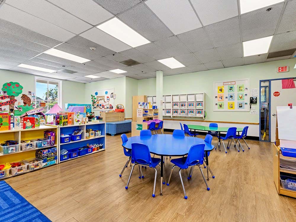 Classroom with blue tables and chairs, shelves of toys and books, and artwork on walls