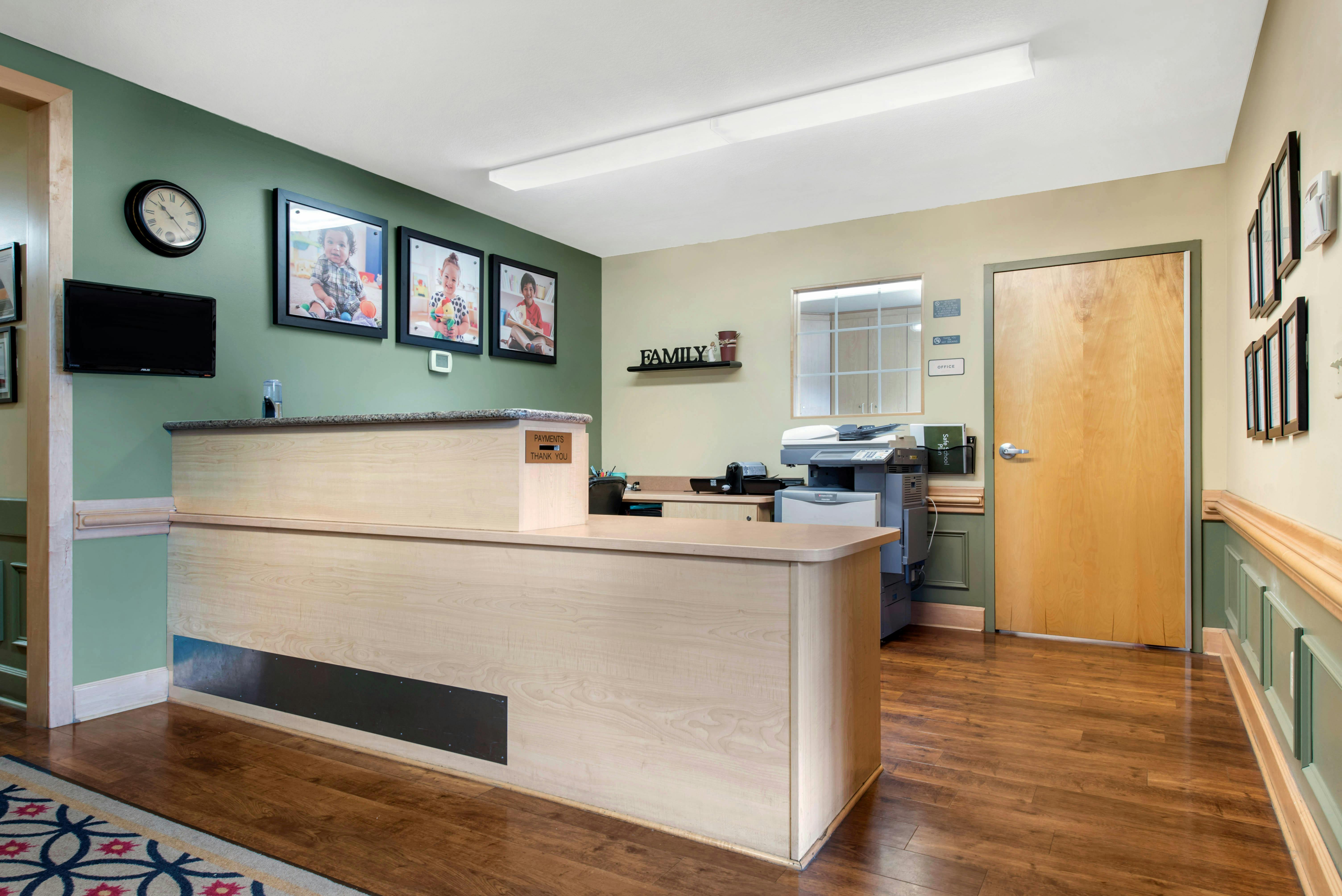 Reception desk with office door, wall-mounted clock, pictures of children, and copier