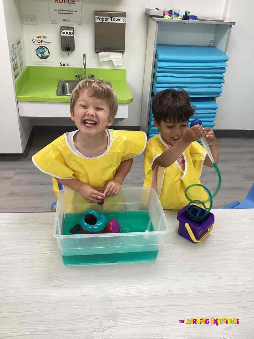 Two children in yellow smocks playing with water and toys in a classroom