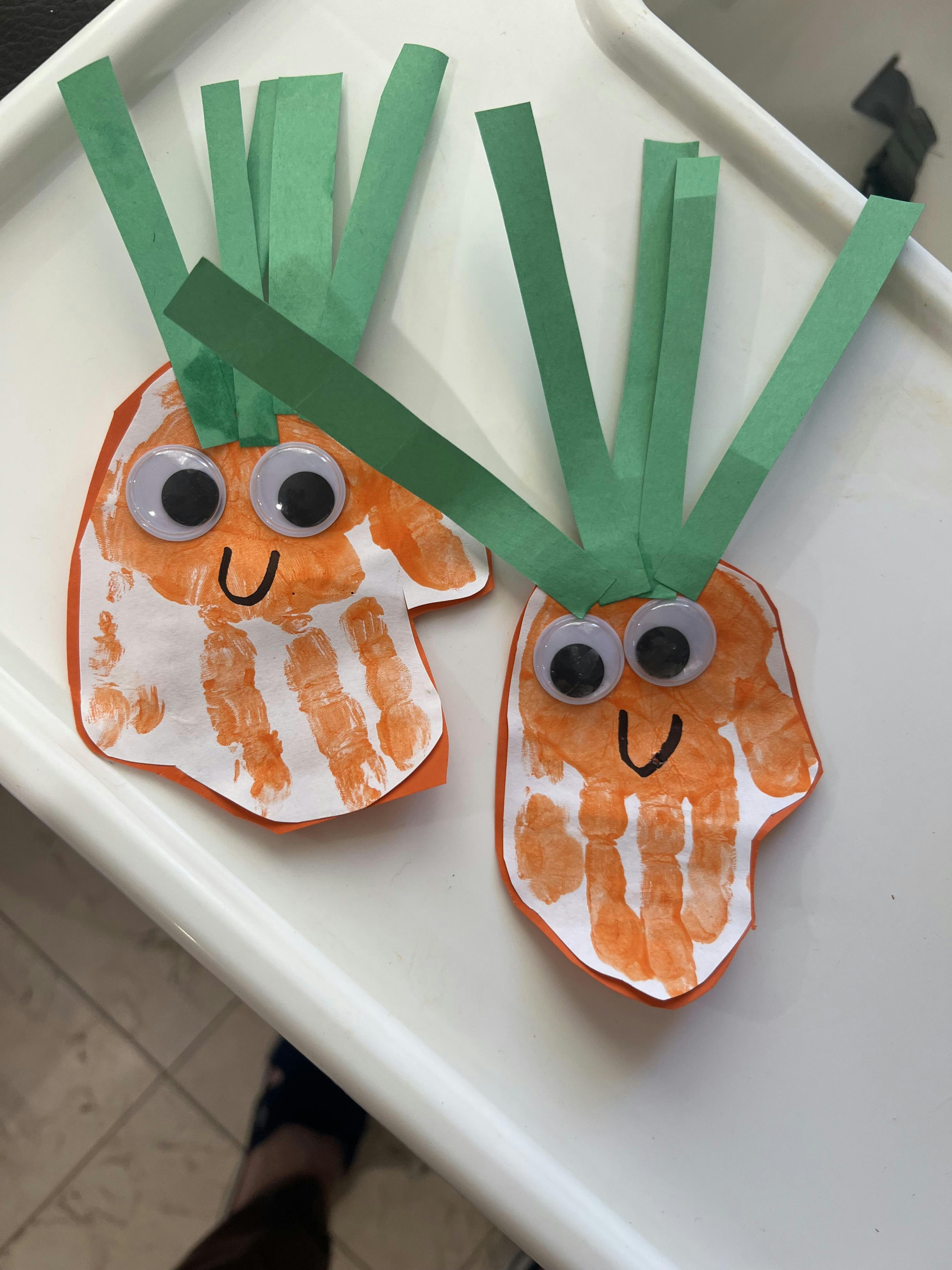 Two paper carrot crafts made with orange handprints and green paper tops