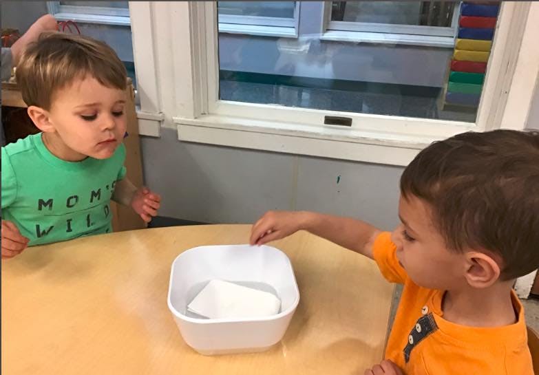 Two children at a table with one child touching an object in a bowl