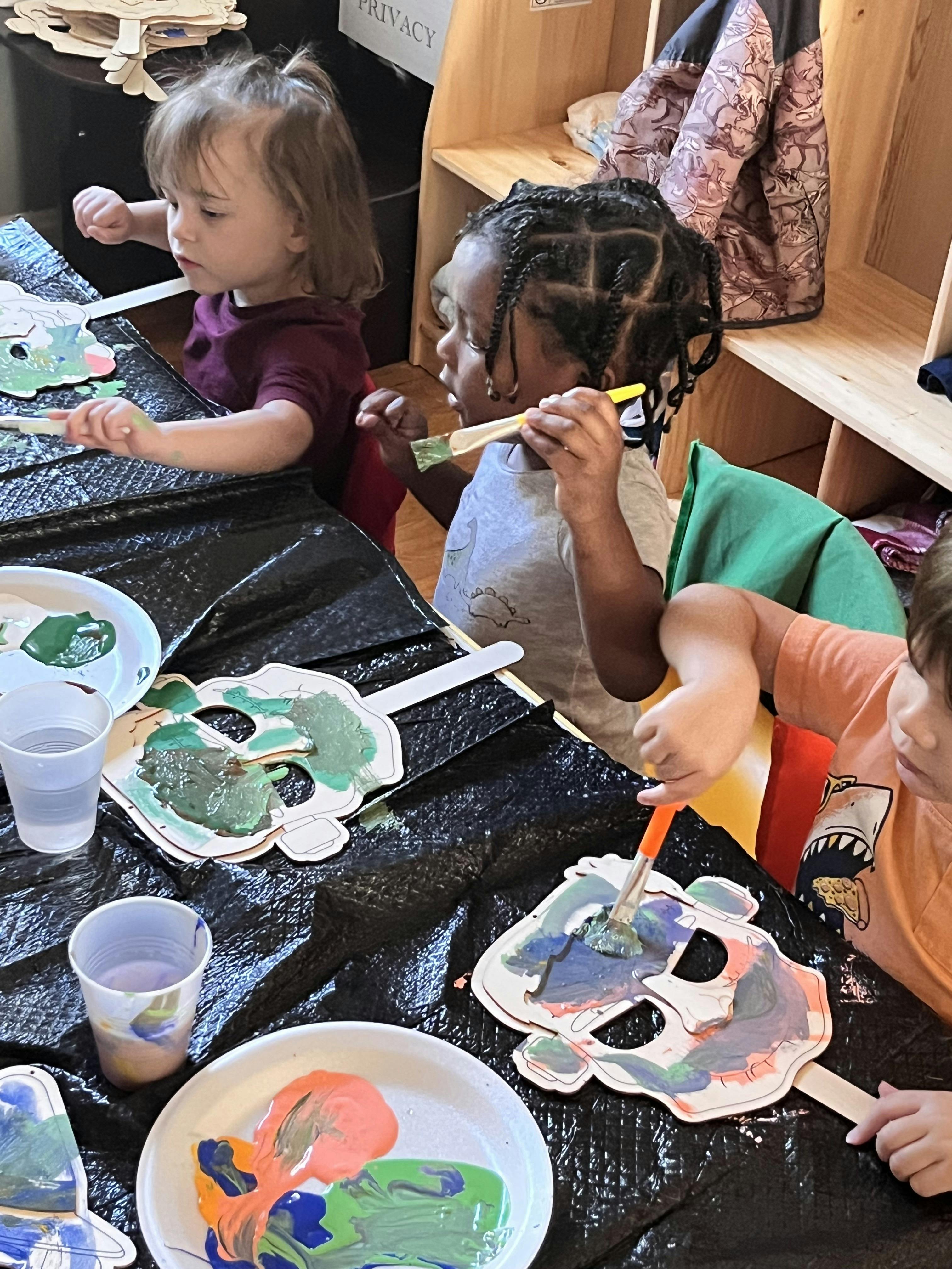 Three children painting paper masks at a table