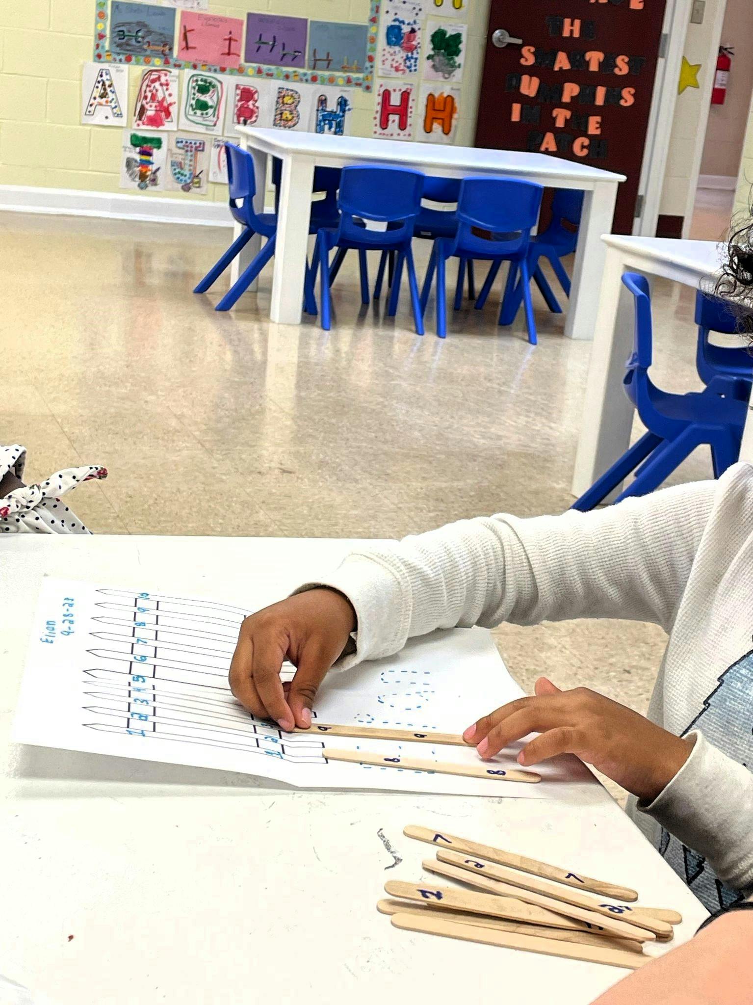 Child's hands working with craft sticks on a paper worksheet in a classroom