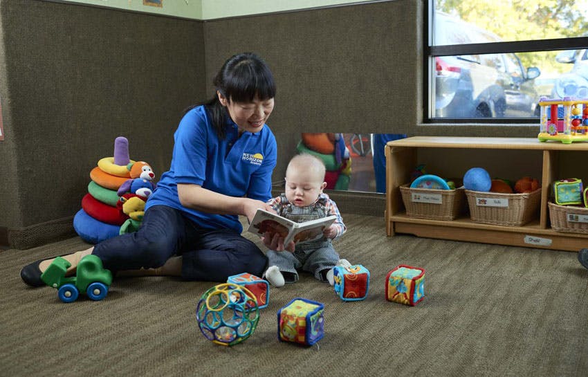 Person reading a book to a baby on the floor surrounded by toys