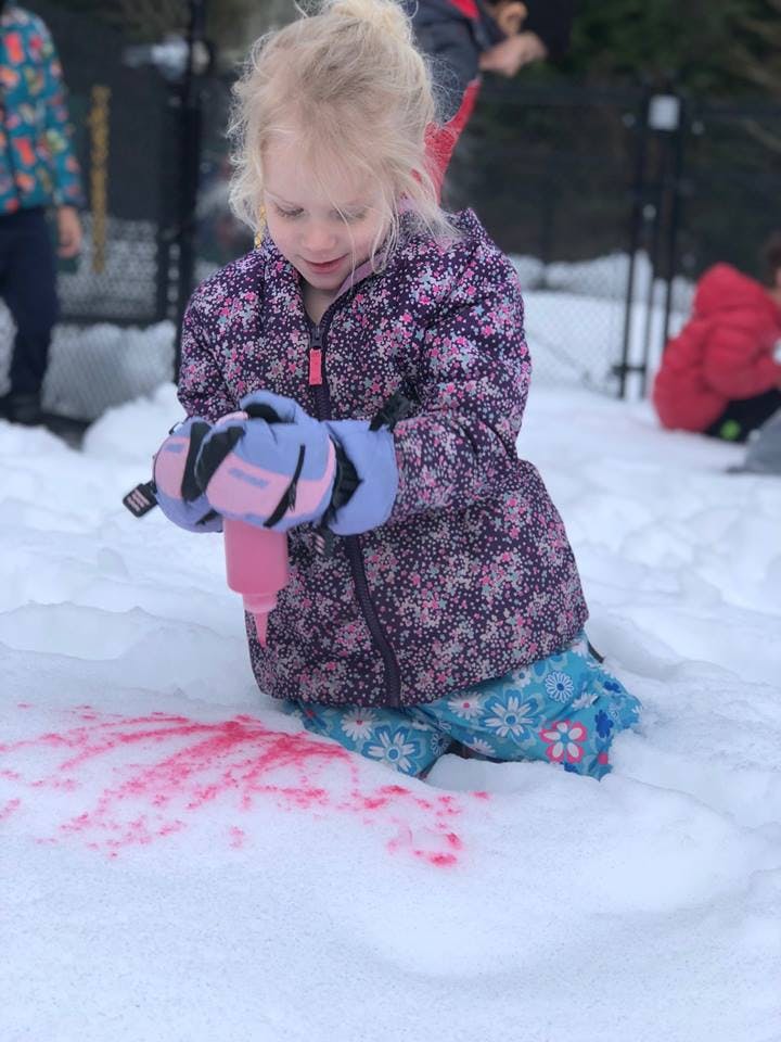 Child in winter clothing drawing on snow with a pink bottle
