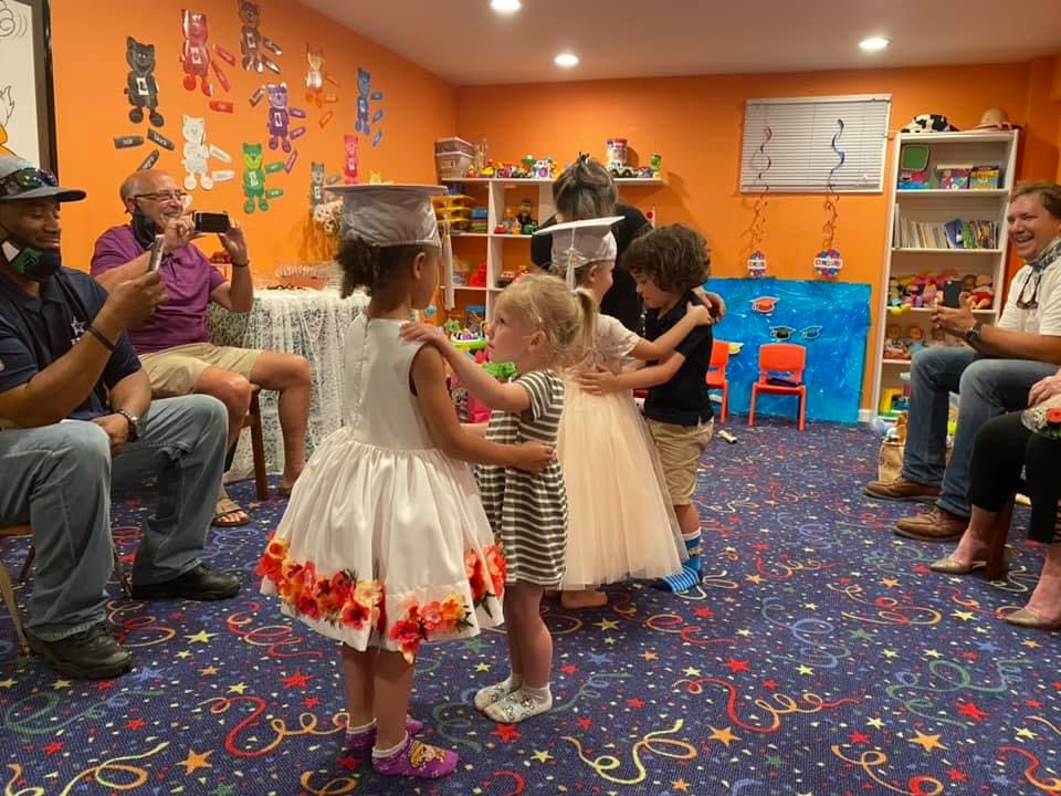 Children in graduation caps and dresses dancing in a room with adults watching