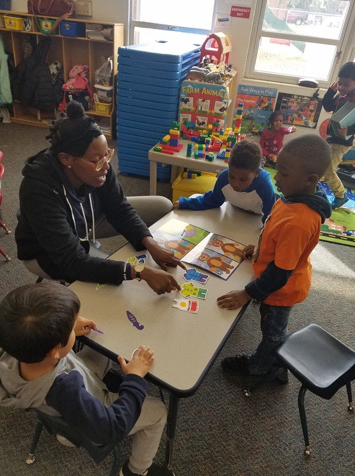 Adult and three children at a table with a book and puzzle pieces in a classroom