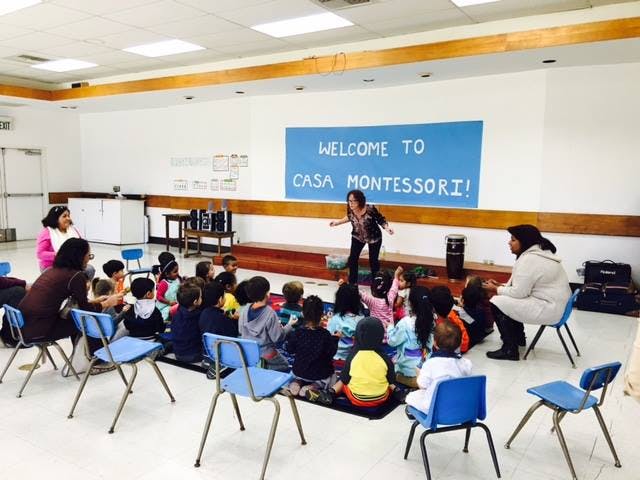 Adult speaking to a group of children and adults in a room with a "WELCOME TO CASA MONTESSORI!" sign