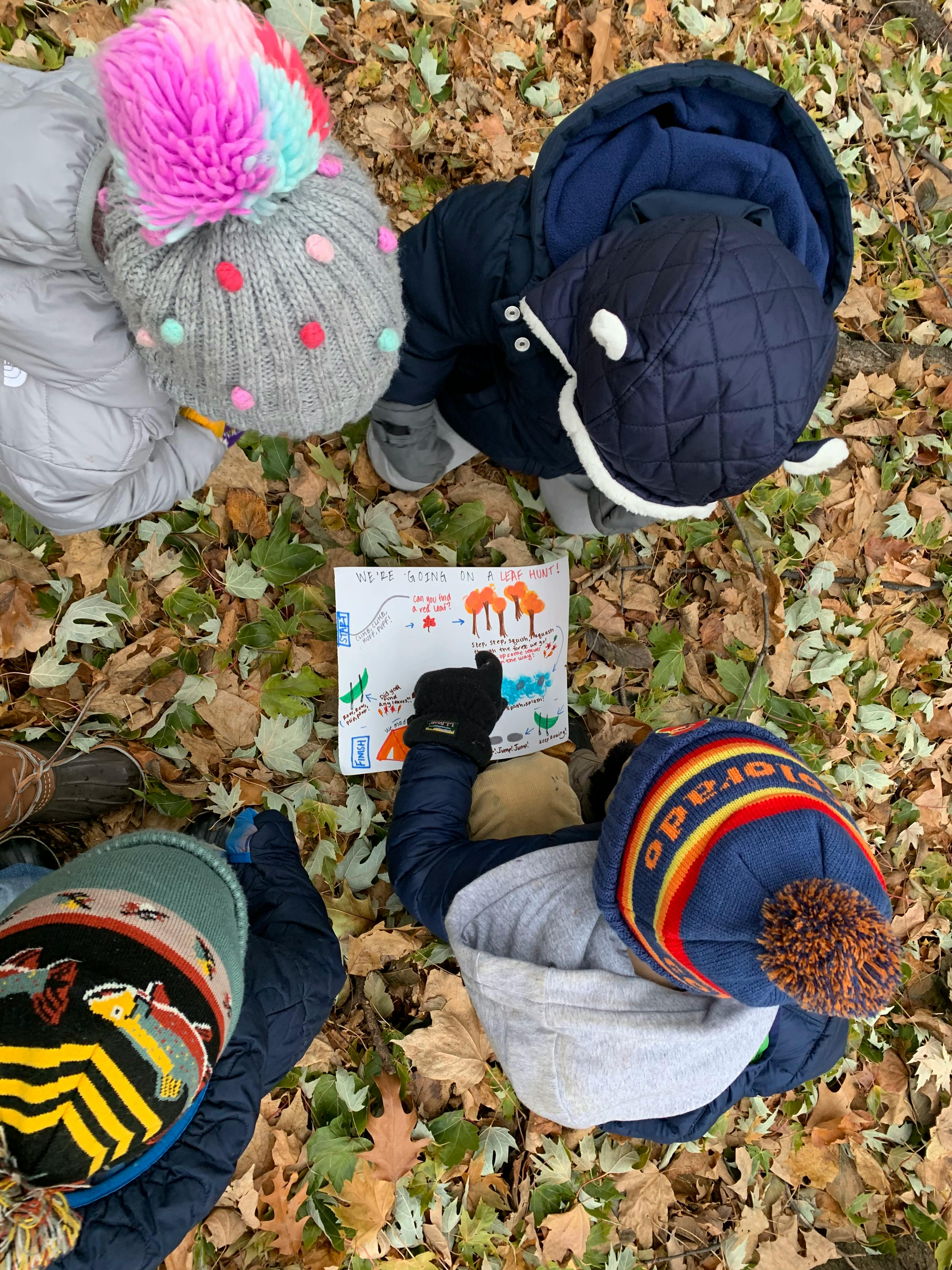 Children on a leaf-covered ground looking at a paper with "We're Going on a Leaf Hunt!"