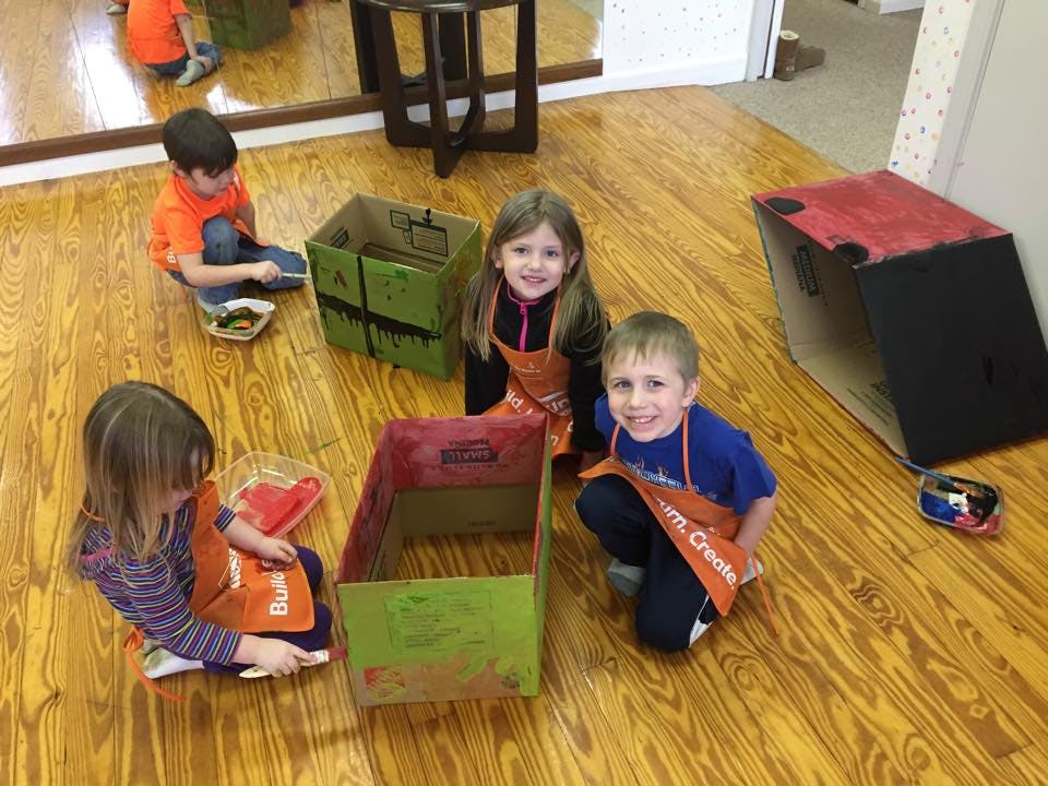 Children in aprons painting cardboard boxes on a wooden floor