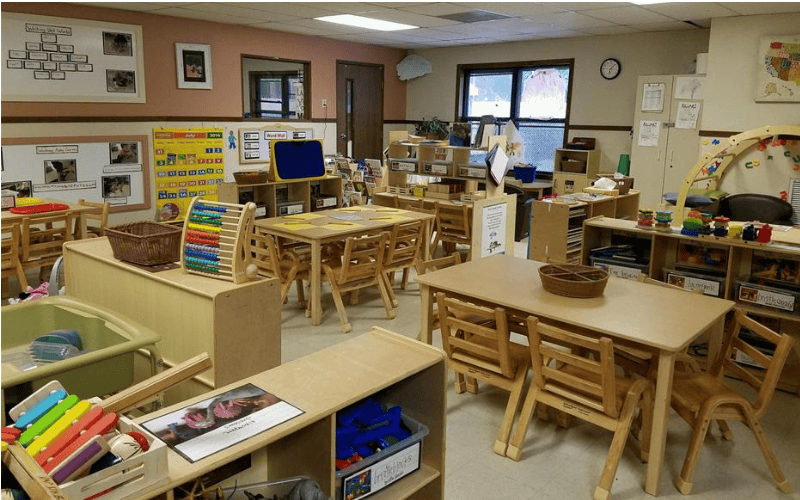 An empty classroom with small wooden tables and chairs, toys, and educational posters