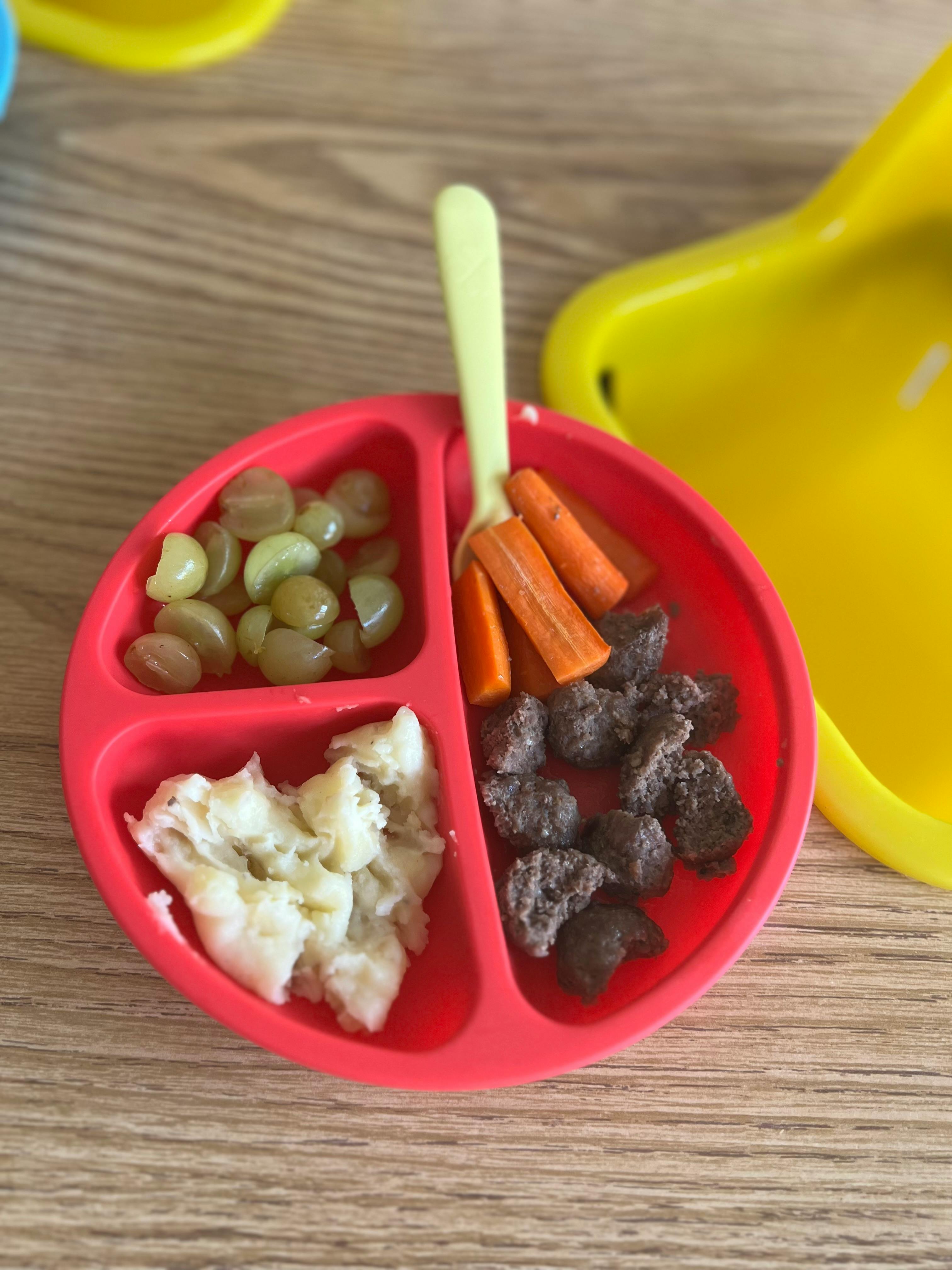 Red divided plate with grapes, carrots, mashed food, and meat