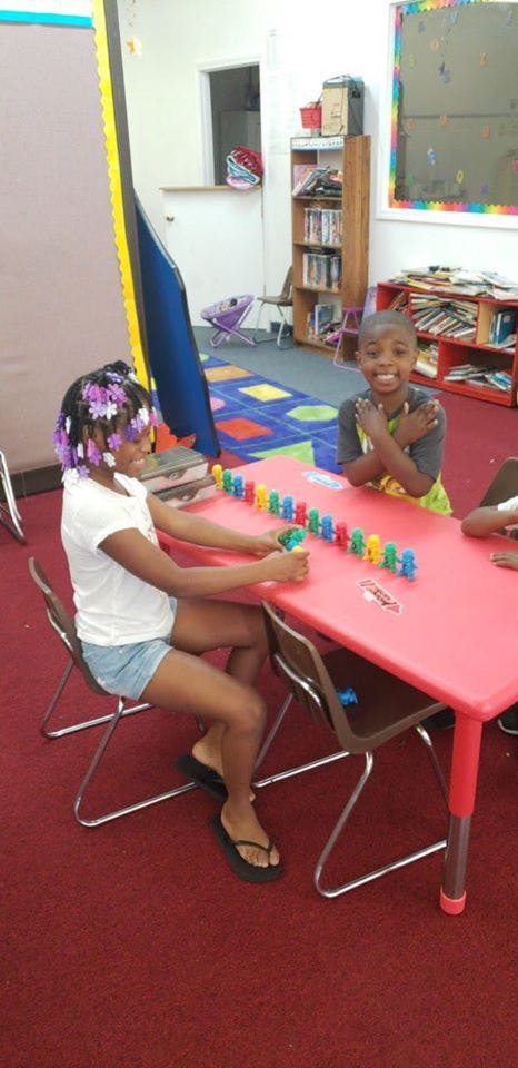 Two children playing with colorful dominoes at a red table in a classroom
