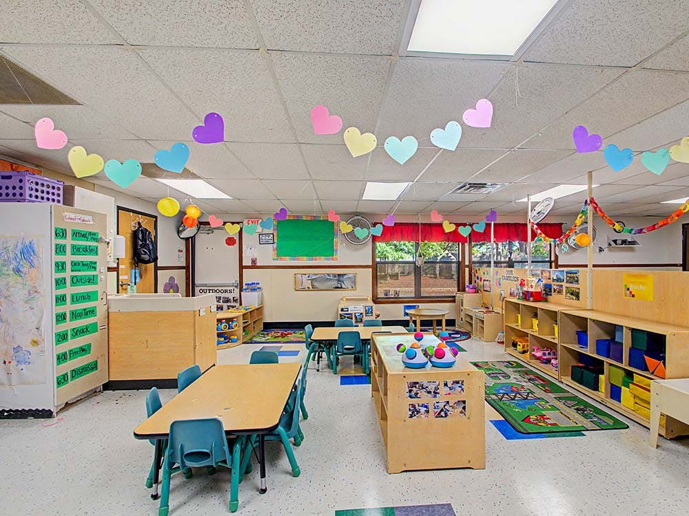 A colorful classroom with small tables and chairs, shelves, and heart decorations
