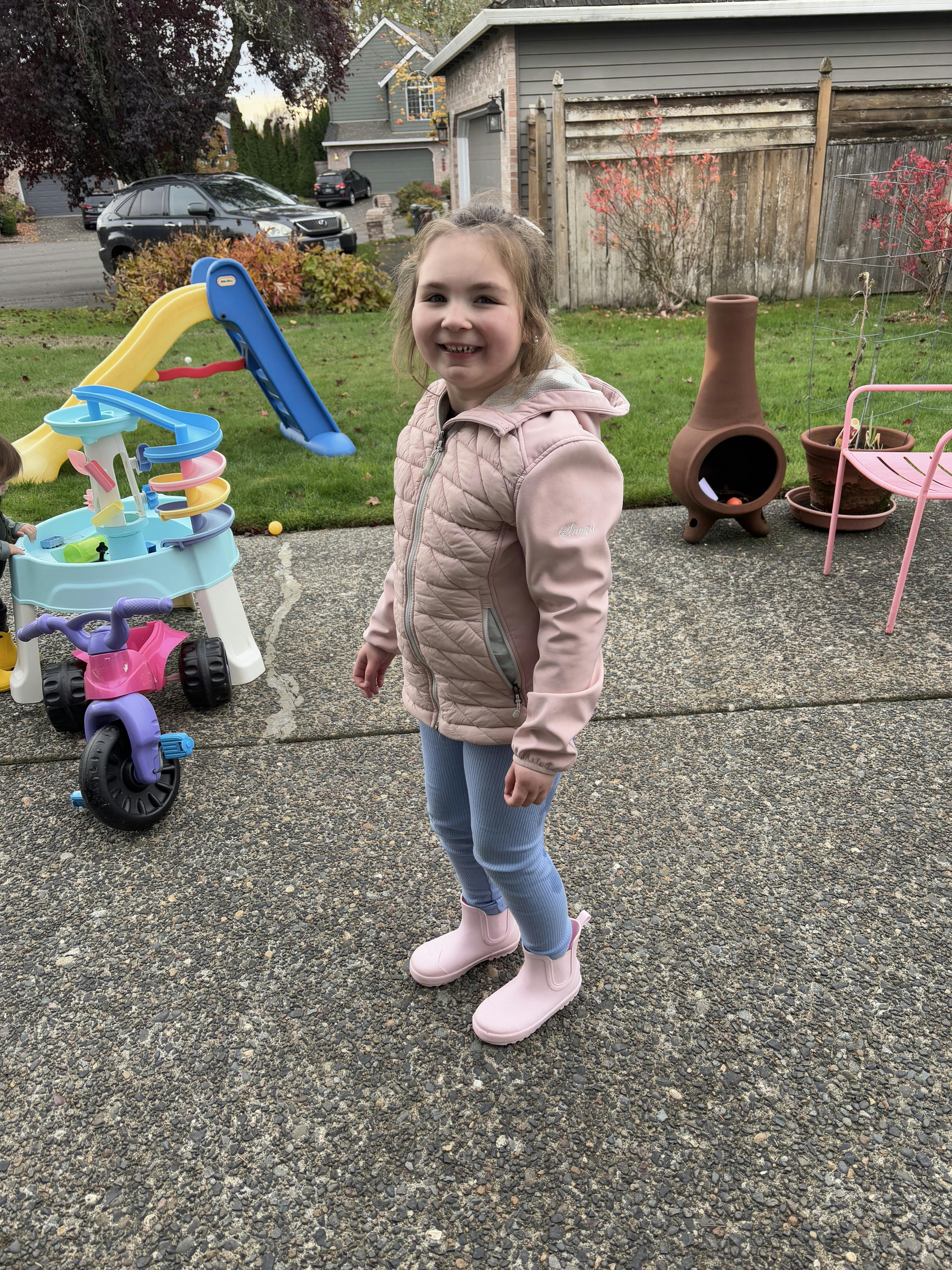 Child in a pink jacket and light blue pants smiling outdoors