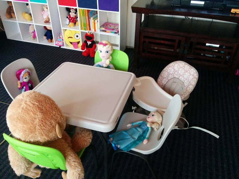 Stuffed animals and dolls sitting around a small table with chairs