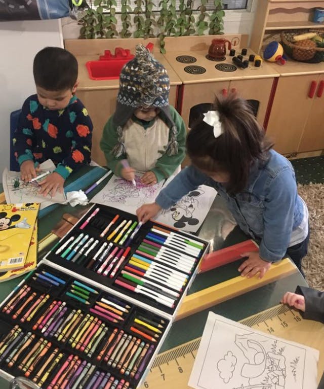Three children coloring at a table with a large art supply box