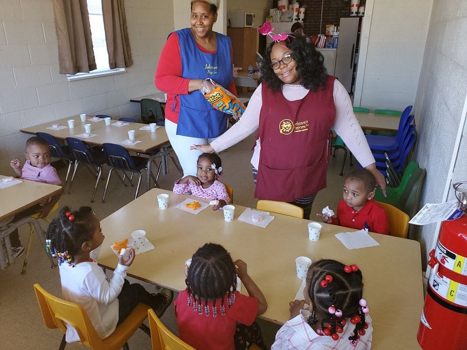 Two people in aprons standing over children seated at tables in a classroom