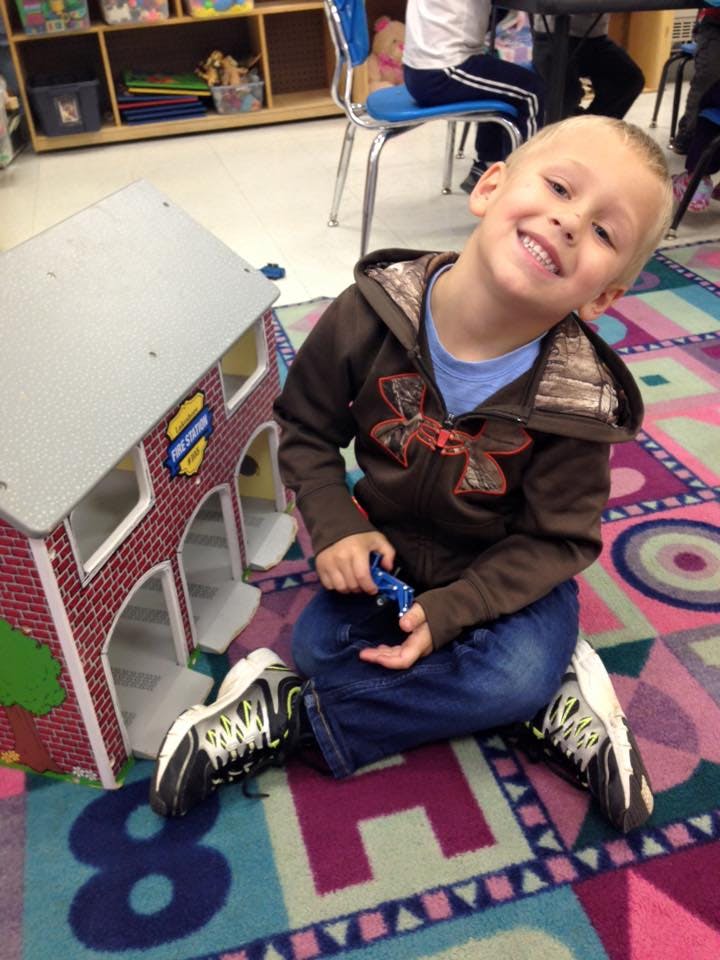 Child sitting on a colorful rug next to a toy house, holding a small toy