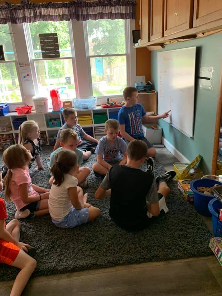 Children sitting on a rug in a classroom, one child writing on a whiteboard