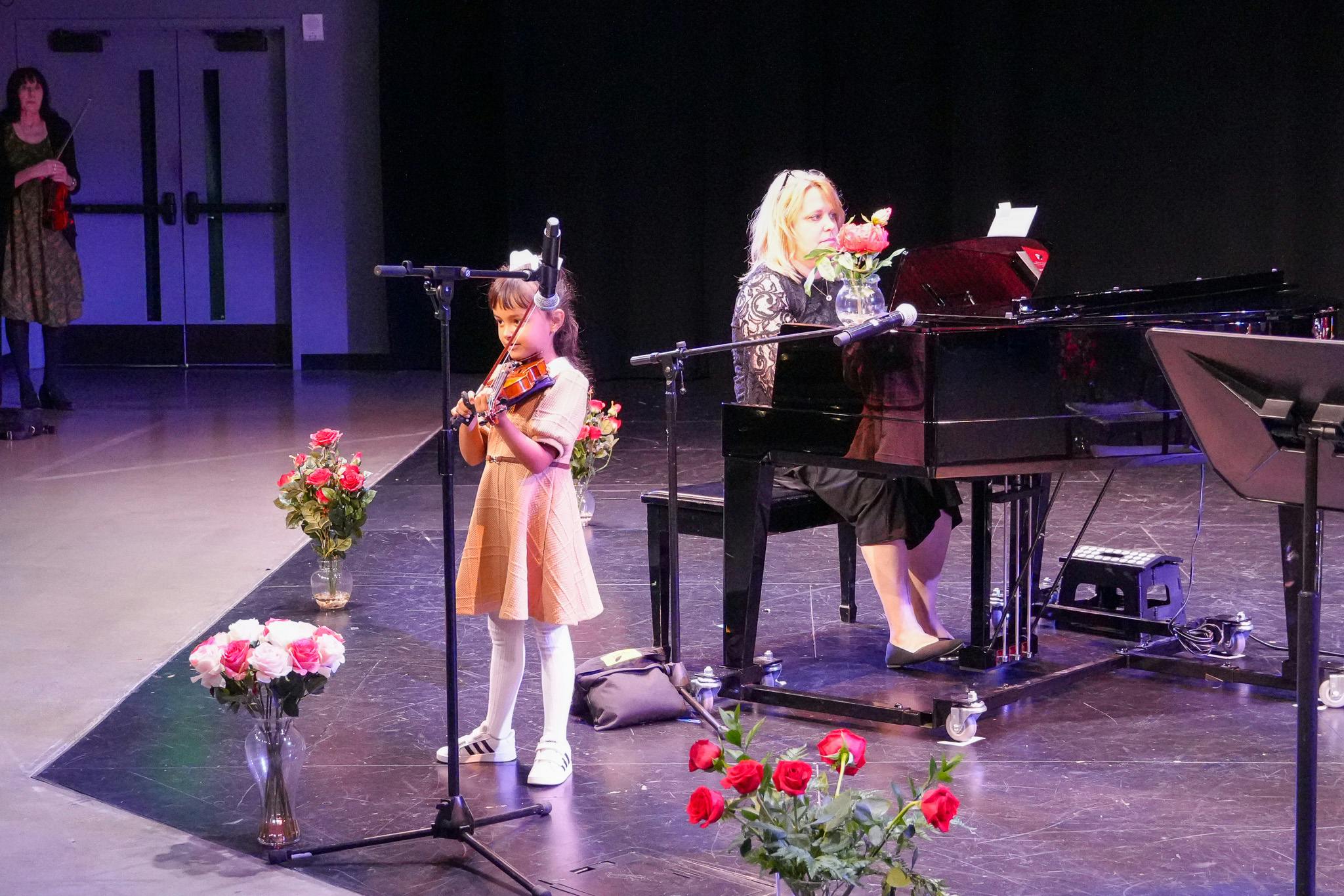 Child playing violin while a person plays piano on a stage