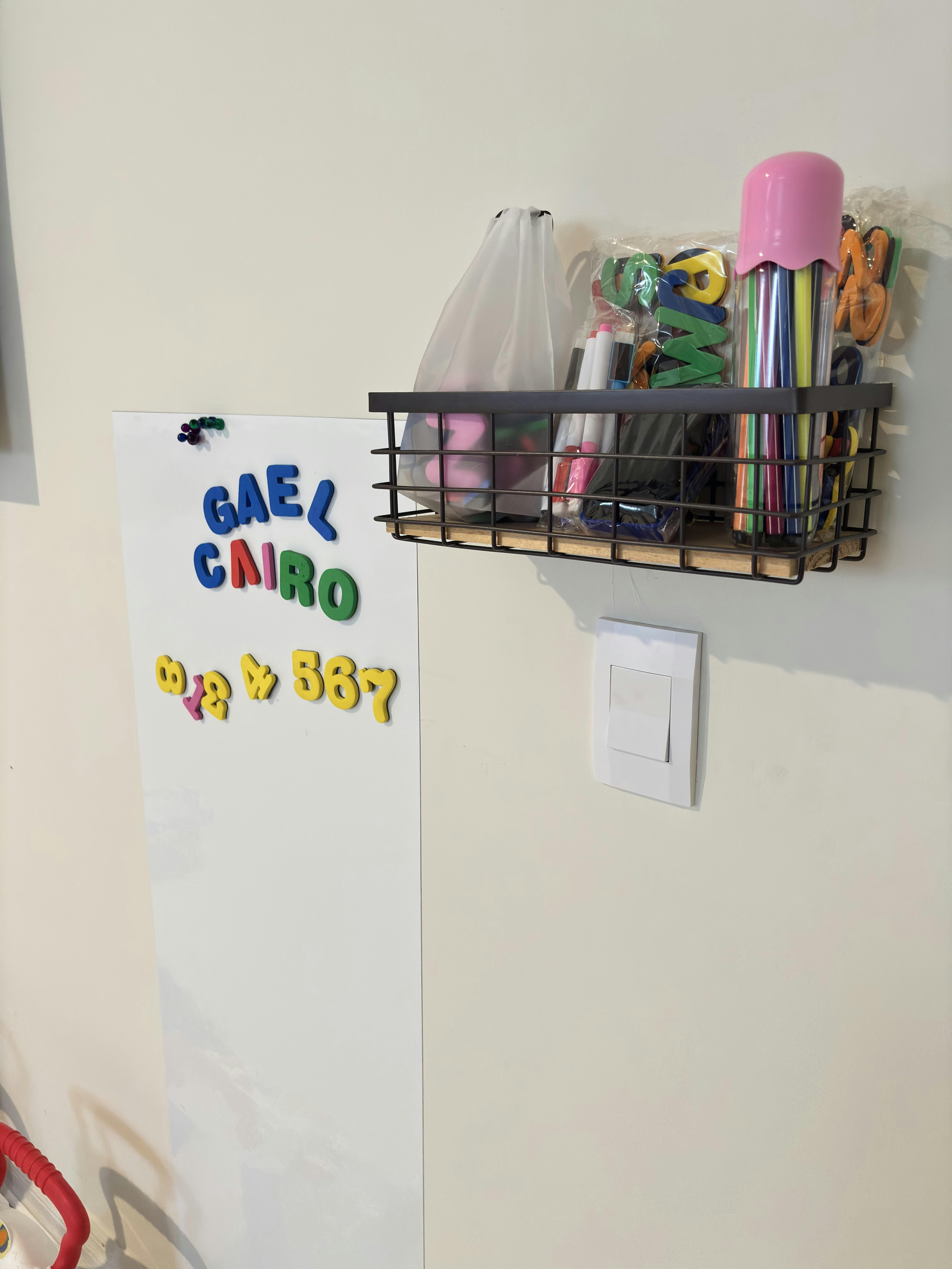 Wall-mounted wire basket holding art supplies next to a whiteboard with magnetic letters