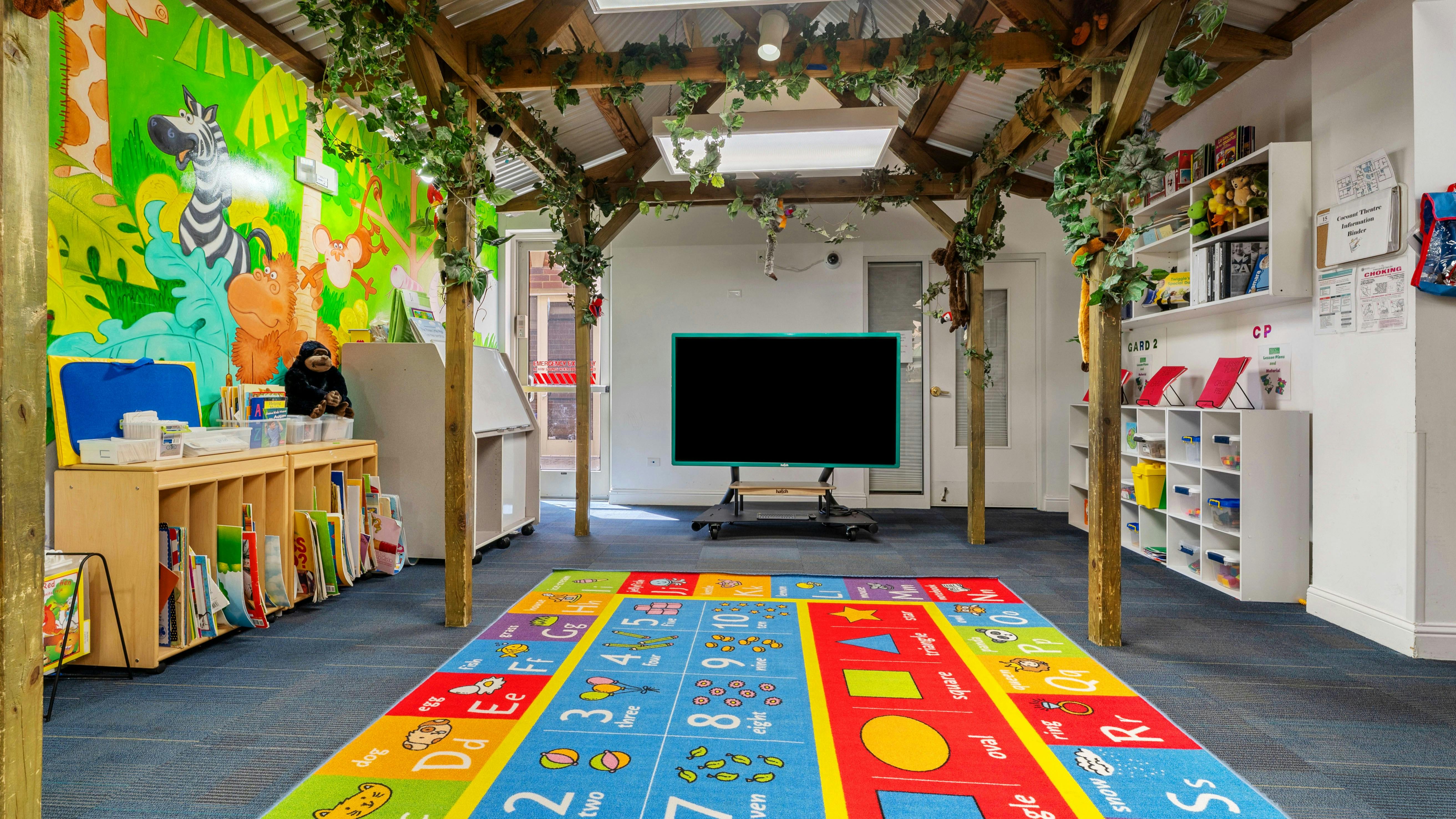 Classroom with jungle-themed mural, colorful alphabet rug, shelves, and a TV