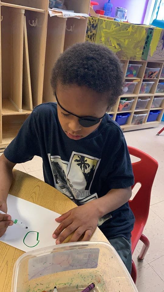 Child with an eye patch drawing on paper at a desk