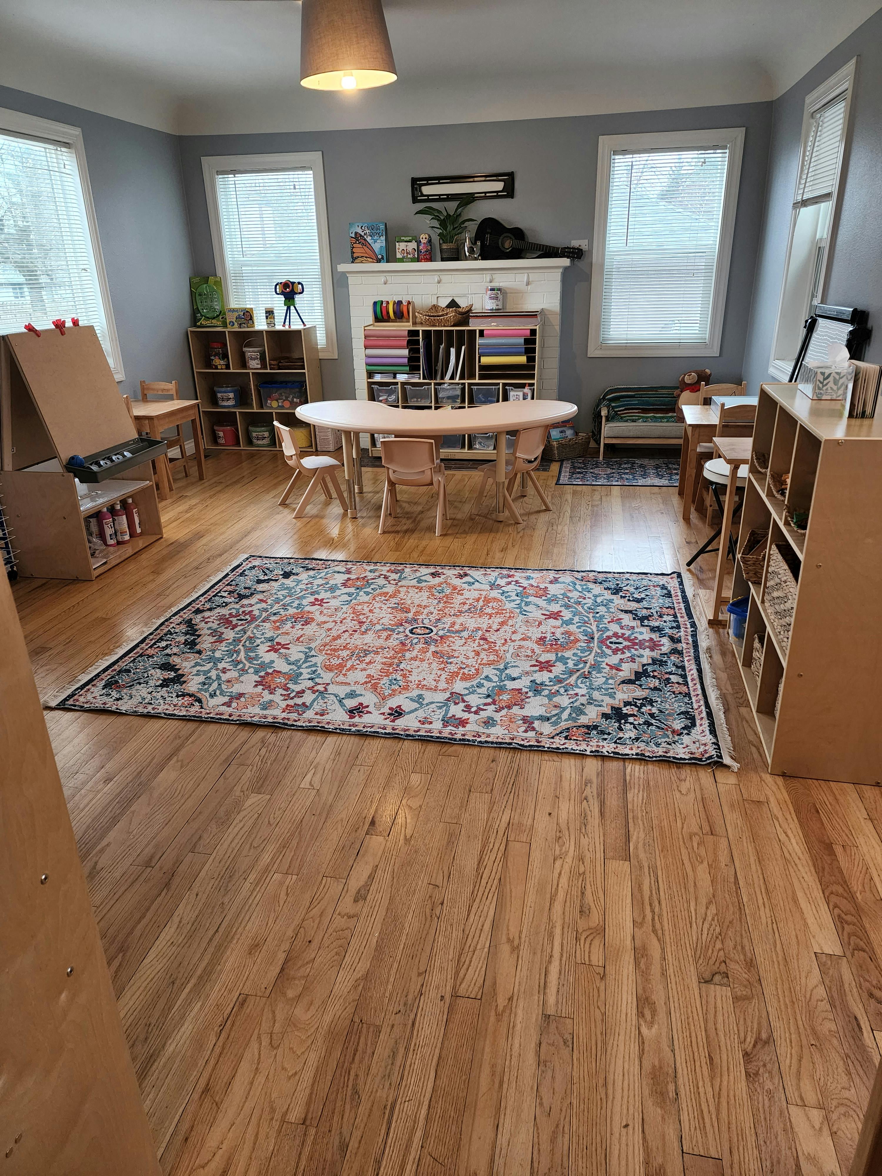 Classroom with tables, chairs, shelves, and a patterned rug on a wooden floor