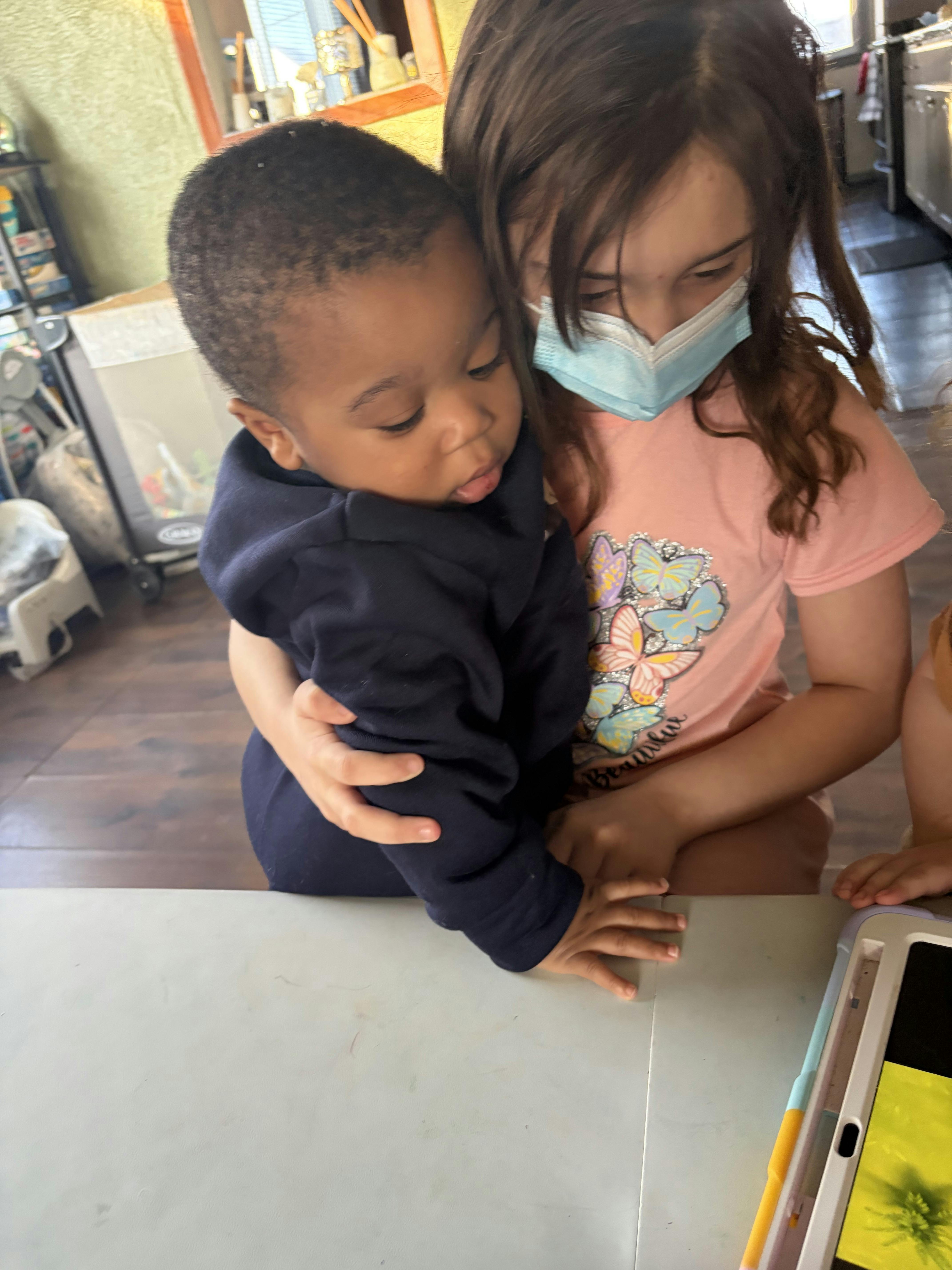 Child wearing a mask holding a baby, looking down at a tablet