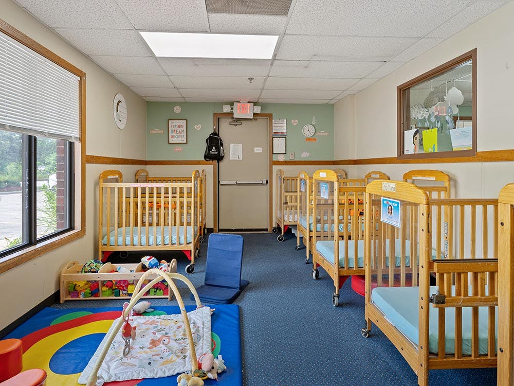 A room with multiple wooden cribs, a playmat on the floor, and a window
