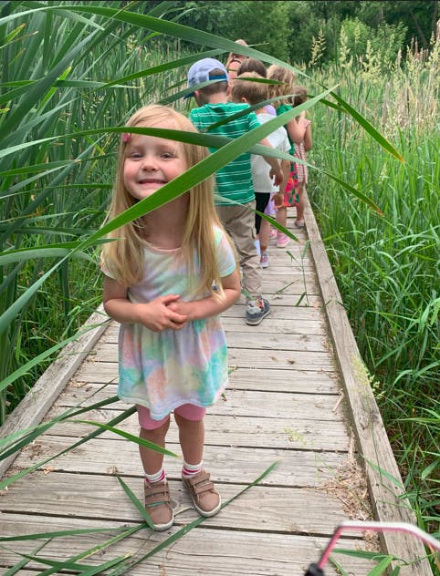 Child on a boardwalk with tall grass, other children in line behind