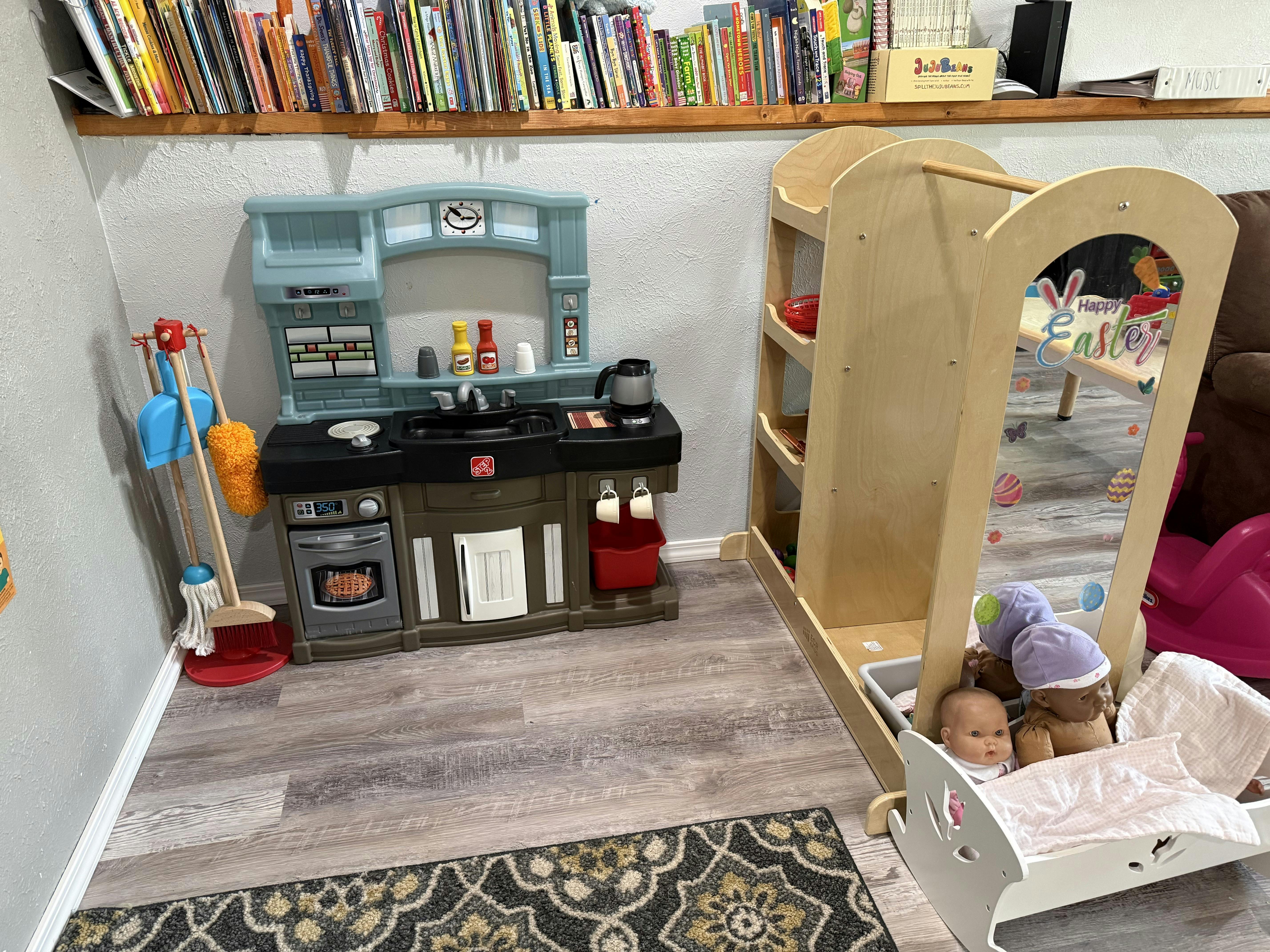 Child's play area with a toy kitchen, cleaning set, mirror, and doll bed with dolls