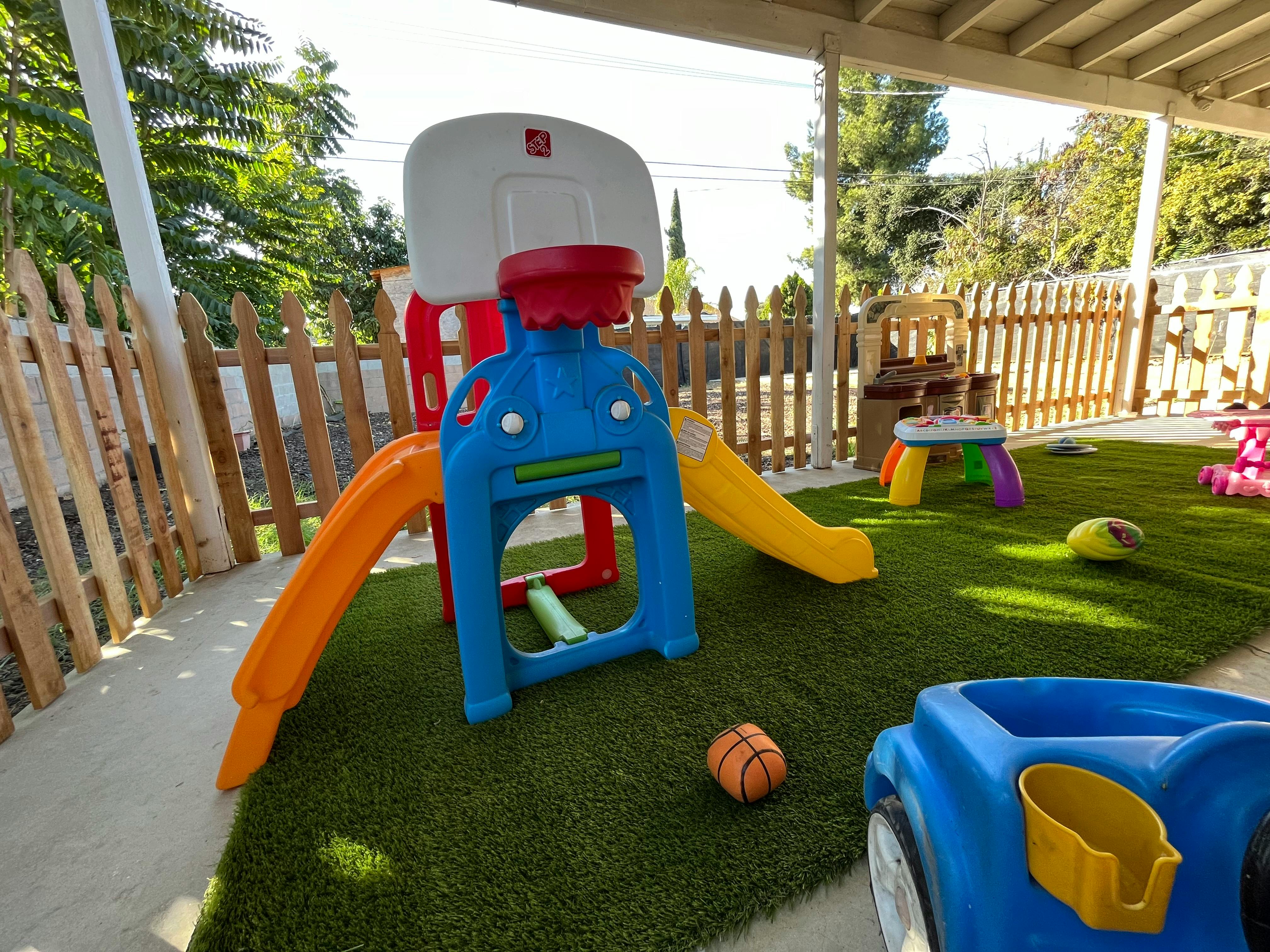 Outdoor play area with a small basketball hoop, slides, and toys on artificial grass