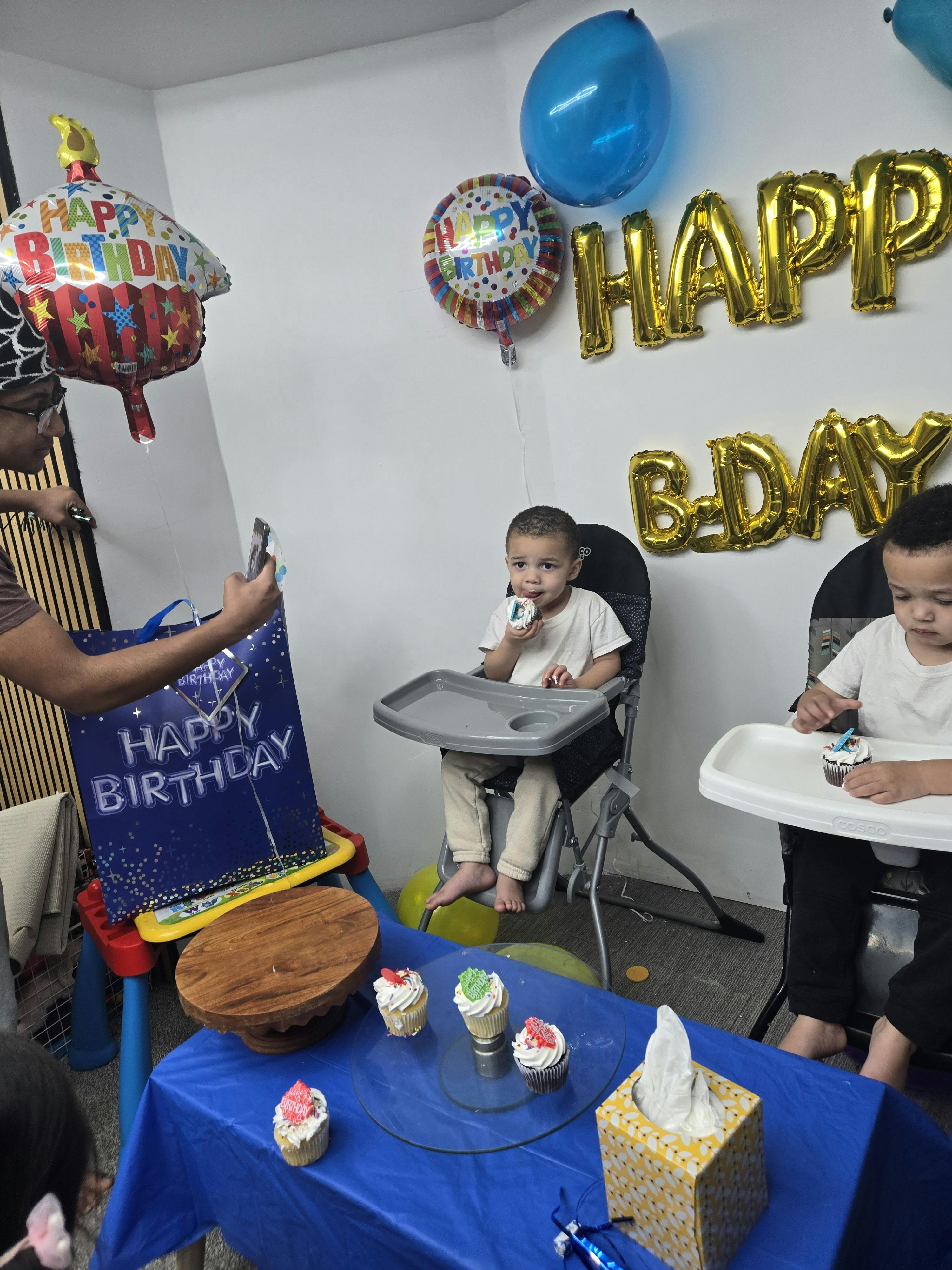 Two children in high chairs with cupcakes, balloons, and a "Happy Birthday" gift bag