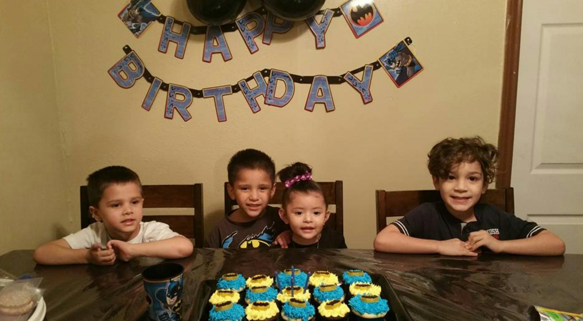 Four children sitting at a table with cupcakes and a "Happy Birthday" banner