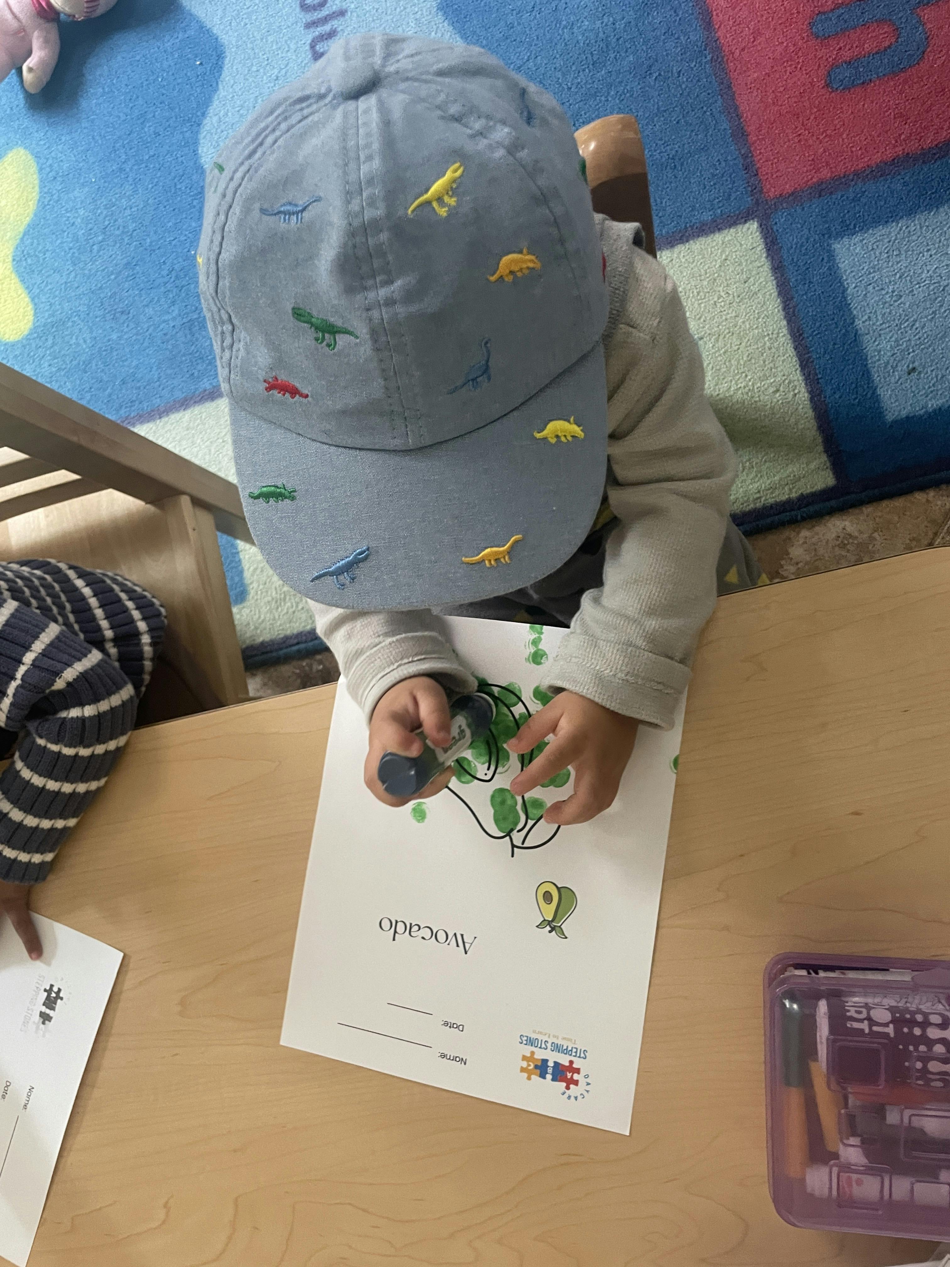 Child wearing a dinosaur hat stamping green dots on an avocado activity sheet