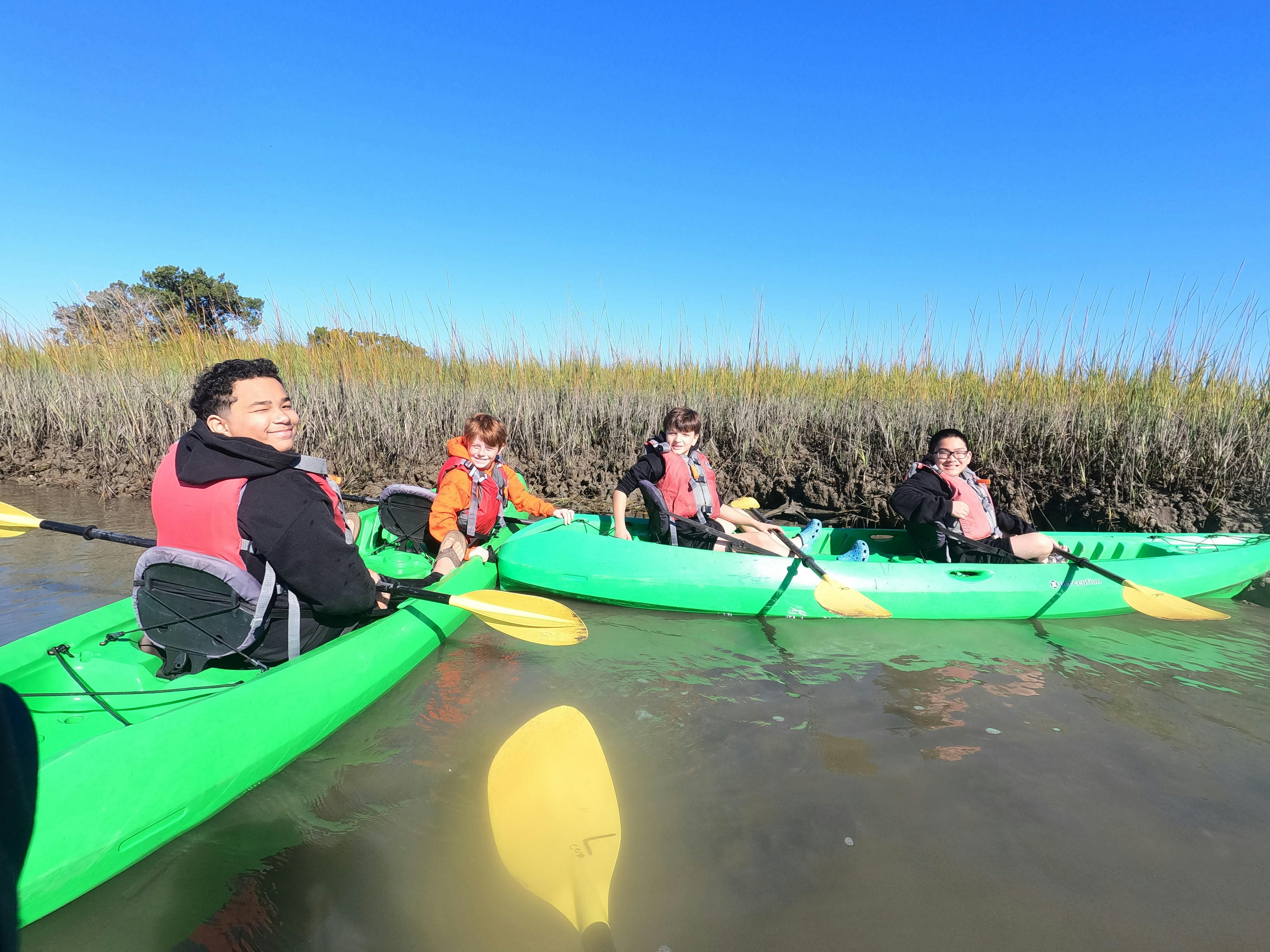 Four people in green kayaks on a sunny day next to marsh grass