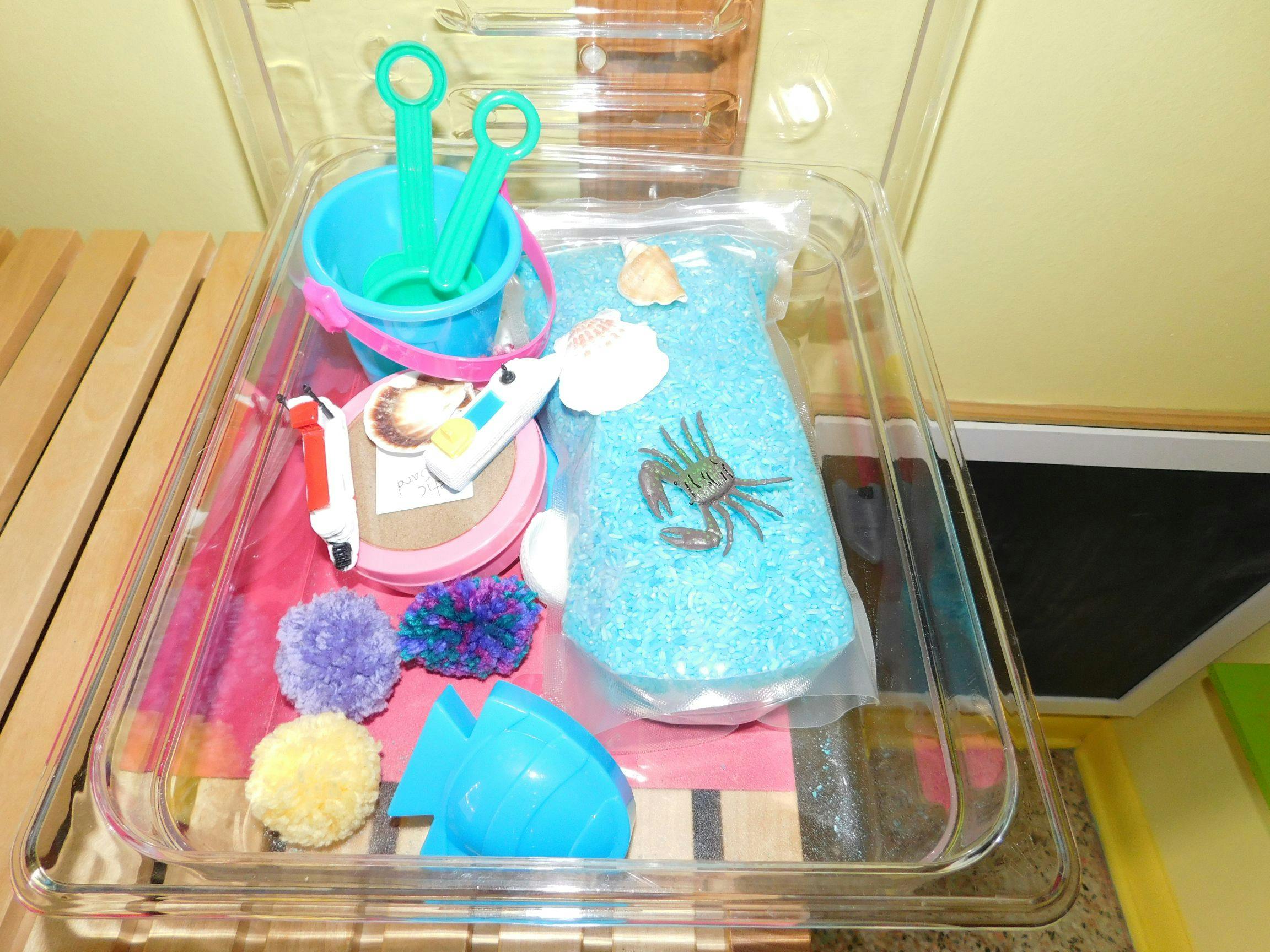 Clear plastic tray with various craft items including pom-poms and sand toys