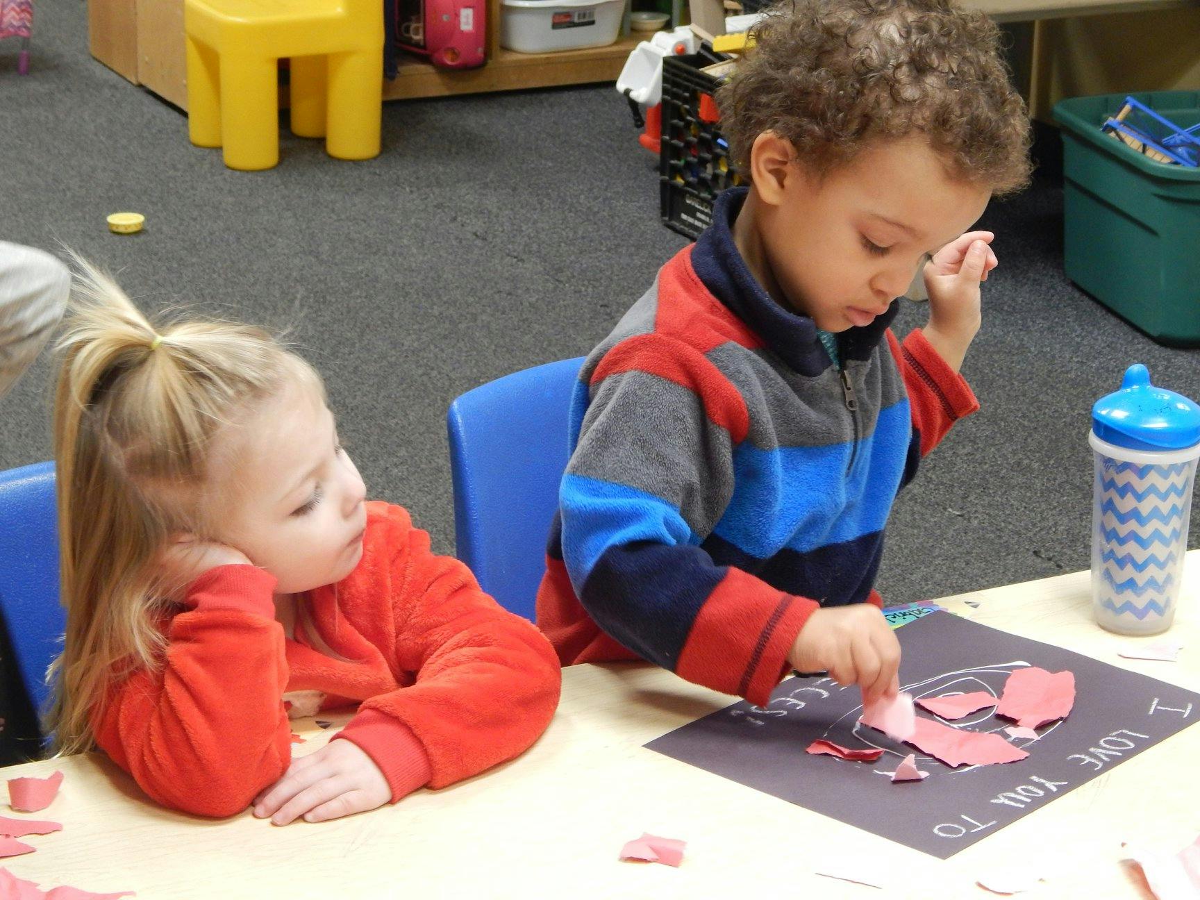 Two children at a table, one working on a craft, the other resting their head